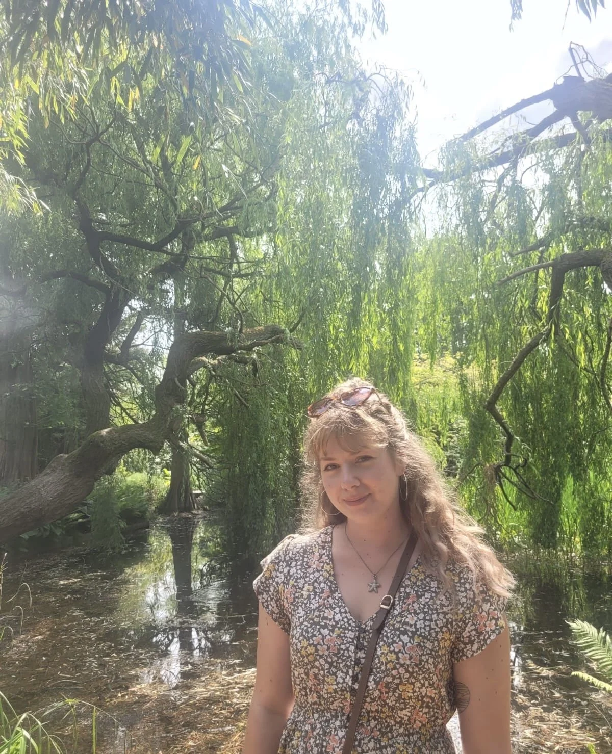 A young woman with curly blonde hair, wearing a floral dress, standing outdoors in front of lush green trees and a small water body, with sunlight filtering through the leaves.