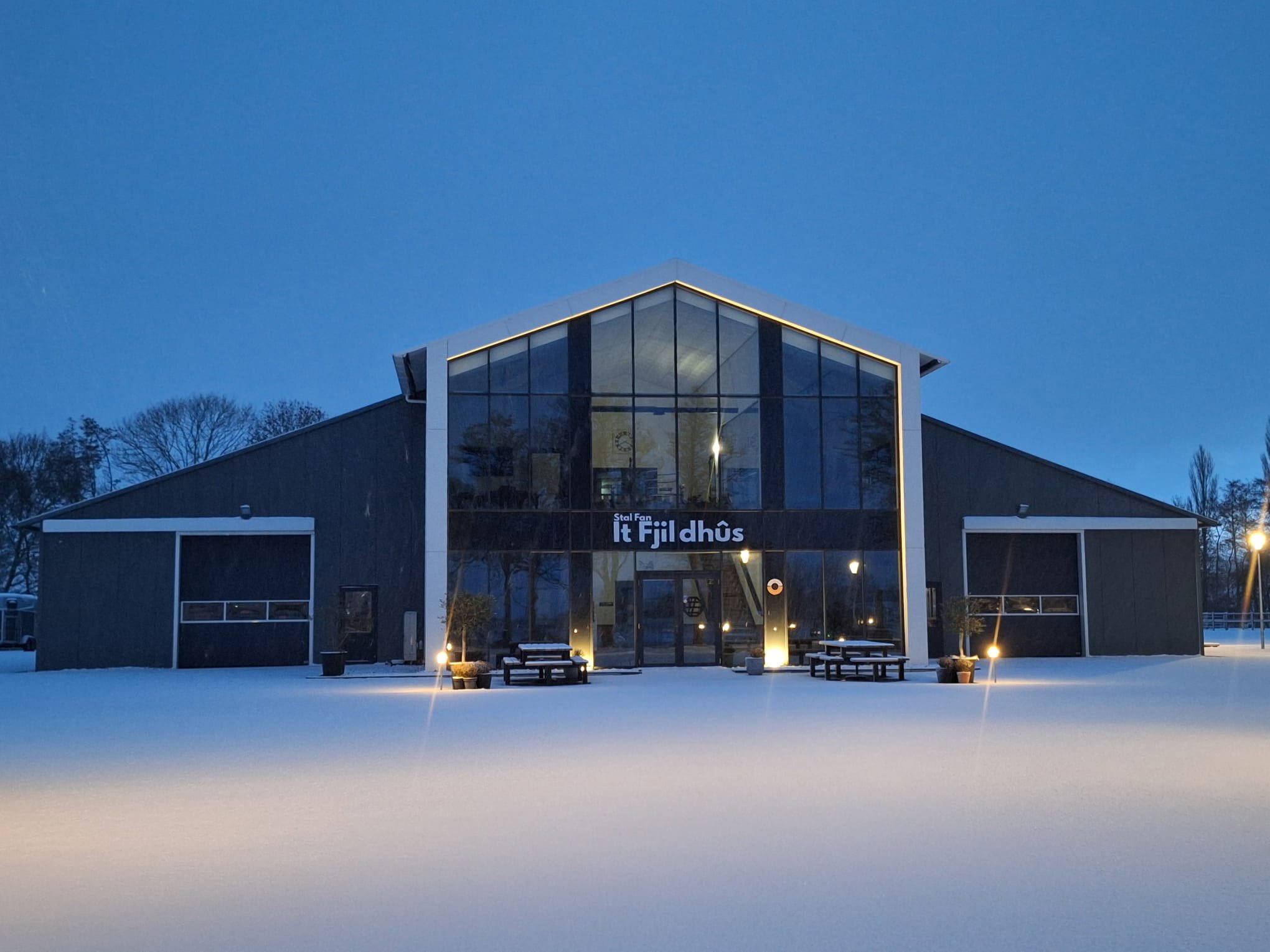 It Fjildhûs in Marsum tijdens een winteravond met verlichte vergaderlocatie en besneeuwd terrein.