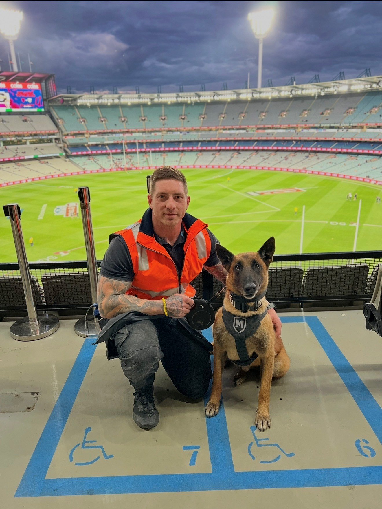 A K9 handler with tattoos and a black shirt wearing an orange safety vest kneeling beside a service dog in a harness at a sports stadium with a large empty field and a dark cloudy sky in the background.