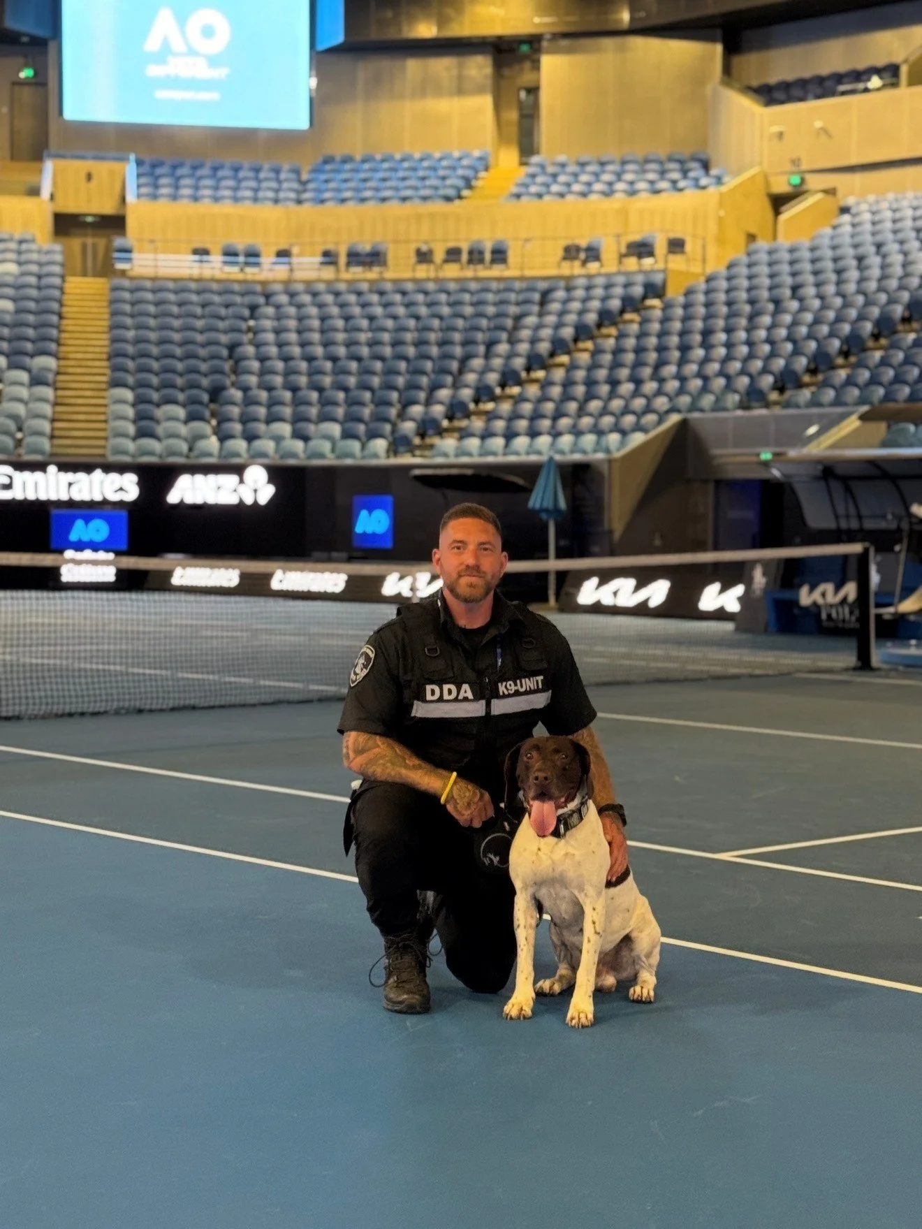 A man in a black tactical uniform with a K9 patch on his chest kneels next to a detection dog sitting on a tennis court inside a stadium. The stadium has empty blue seats and digital advertisements in the background.