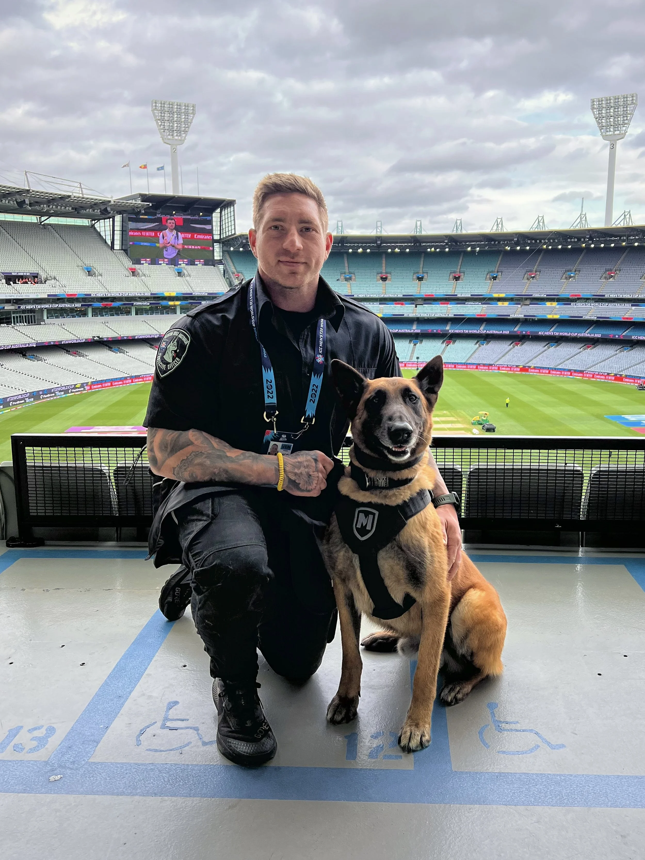A man in police uniform kneeling next to a service dog in a stadium with a view of the field and large screens.