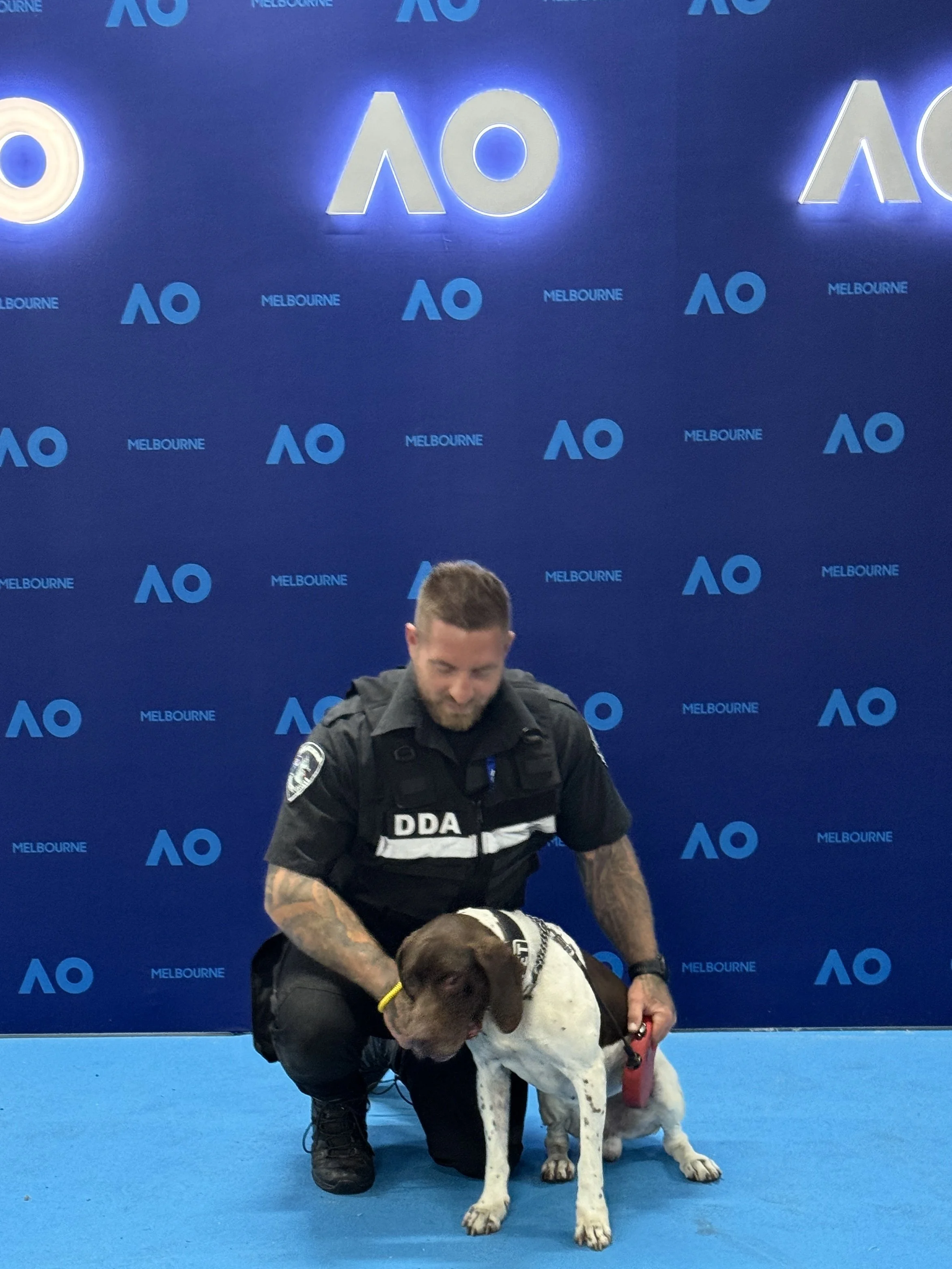 A K9 handler kneeling with a detection dog in front of a blue wall with 'Australian Open' logo.