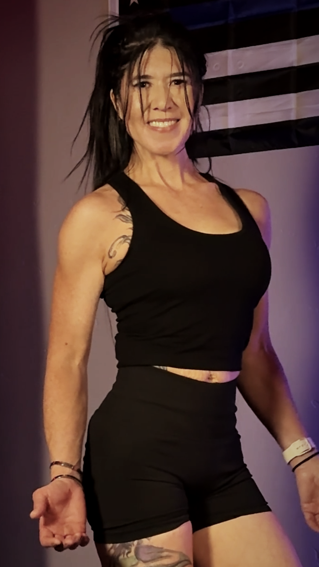 A woman with black hair in a messy style, wearing a black tank top and black shorts, smiling at the camera in an indoor setting with purple and black wall decor.