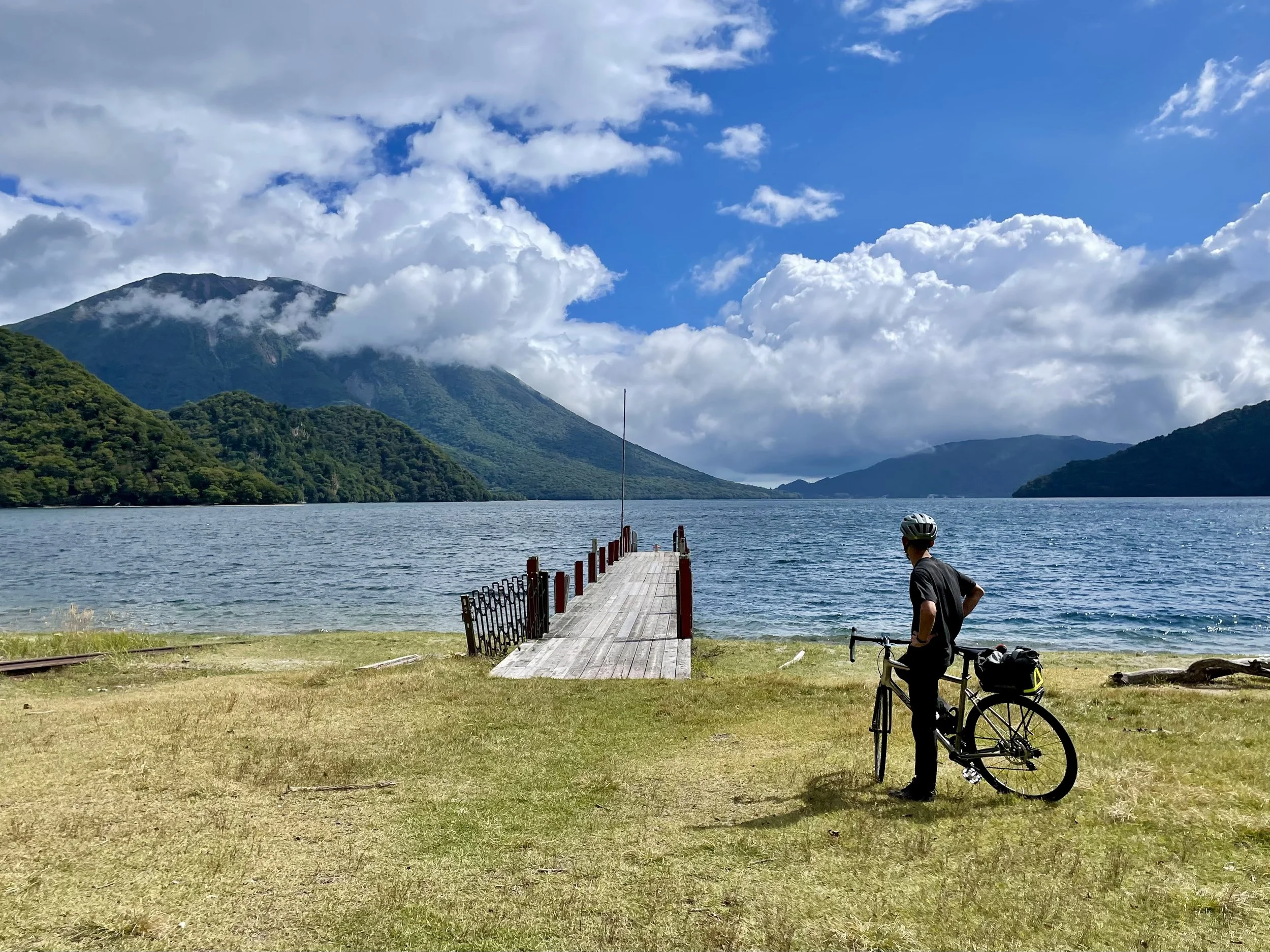 A person wearing a helmet stands next to a bicycle on a grassy shore near a lake, with mountains and partly cloudy sky in the background.