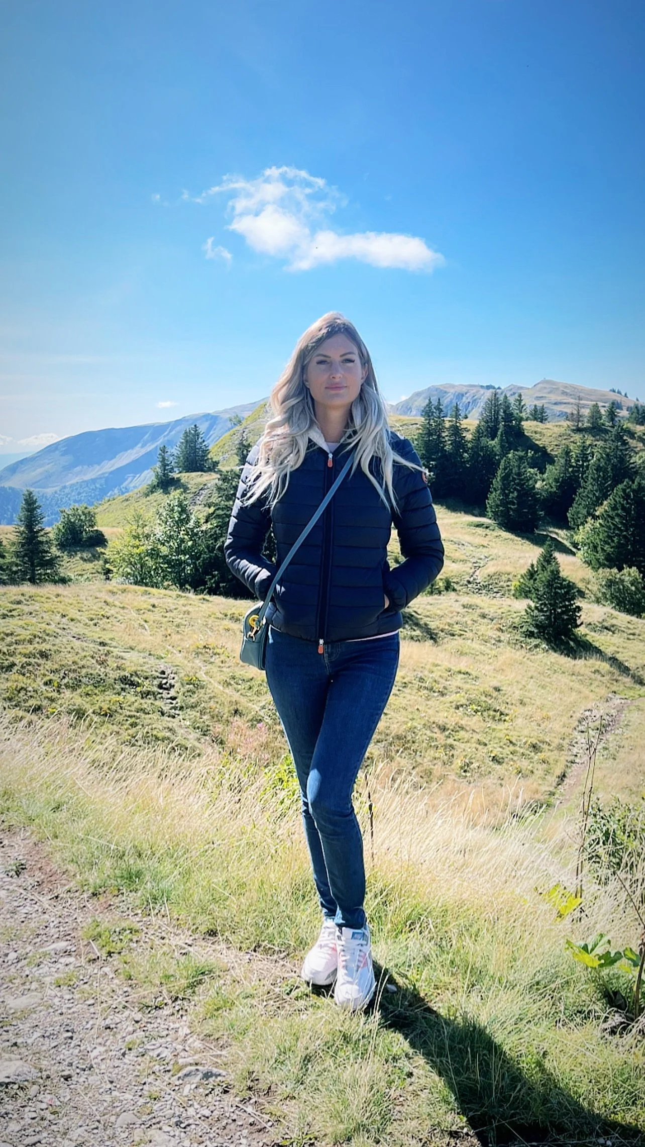 Une femme debout dans un paysage de montagne avec des arbres verts, portant une veste bleu marine, un jean bleu et des baskets blanches, sous un ciel bleu avec quelques nuages.