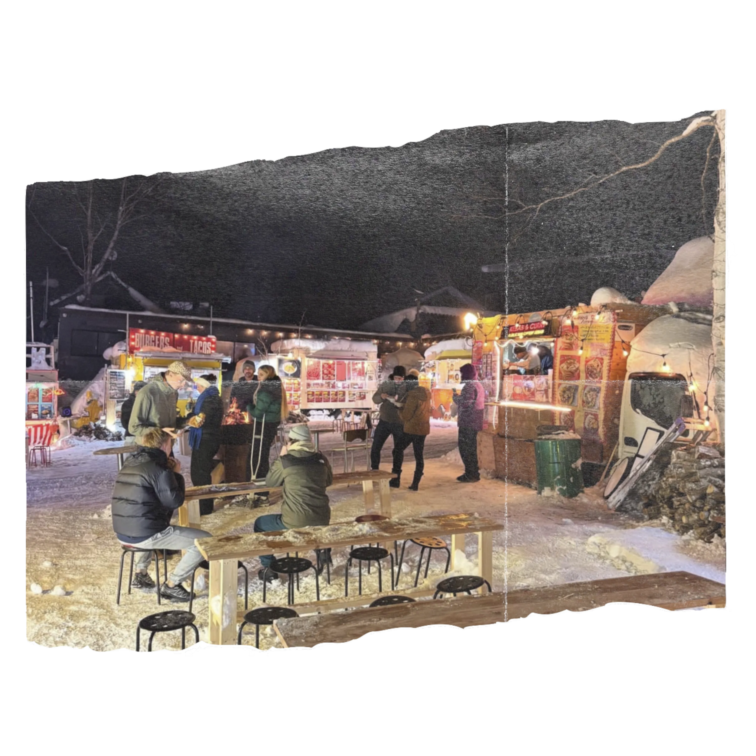 Night scene at an outdoor fair featuring food stalls, people, benches, and tents, with a mountain backdrop under a dark sky.