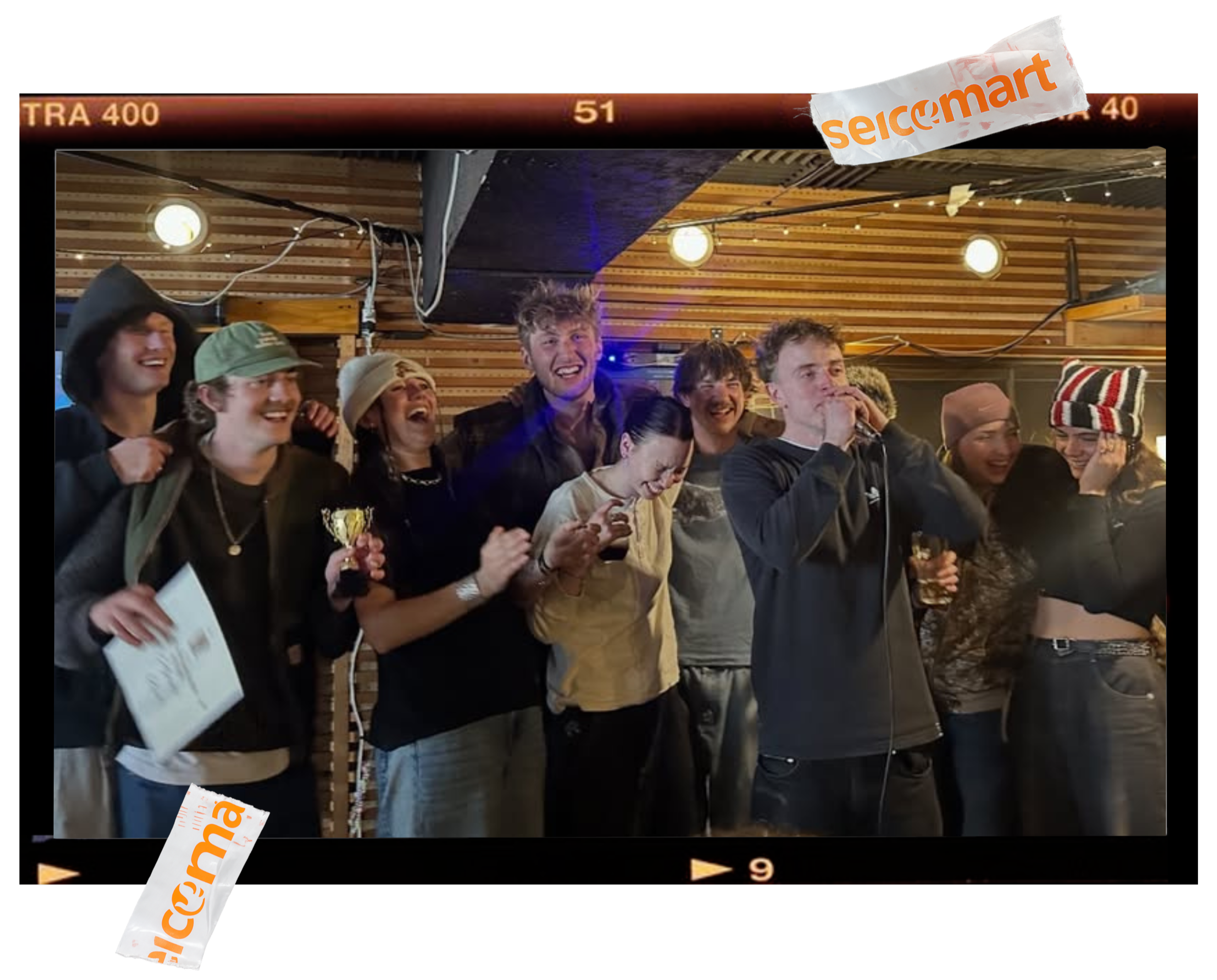 A group of young people smiling and laughing together indoors, some holding drinks, with a background of wooden panels and ceiling lights.