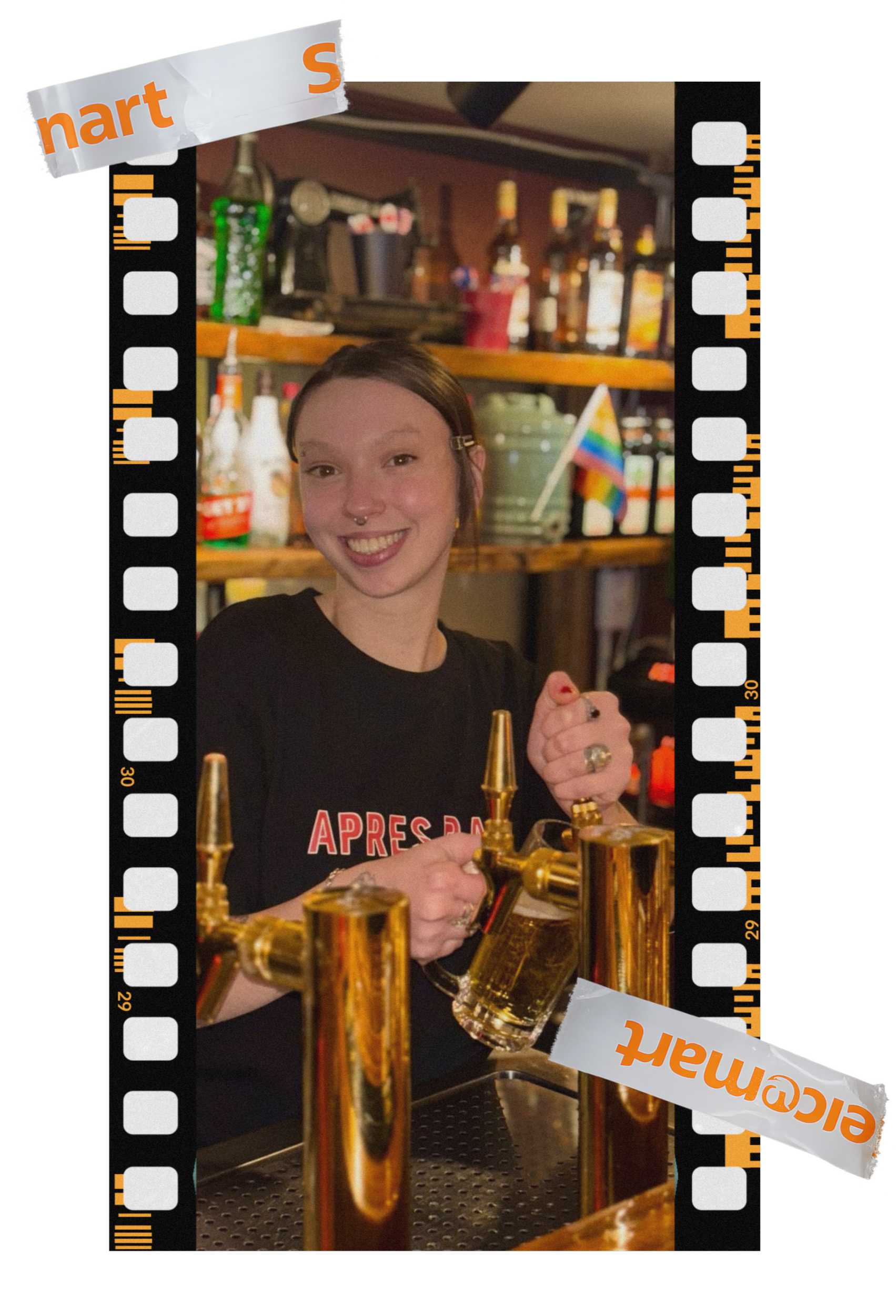 A young woman smiling behind a bar with several beer taps, holding a glass of beer, in a cozy setting with shelves of bottles and a small rainbow pride flag.