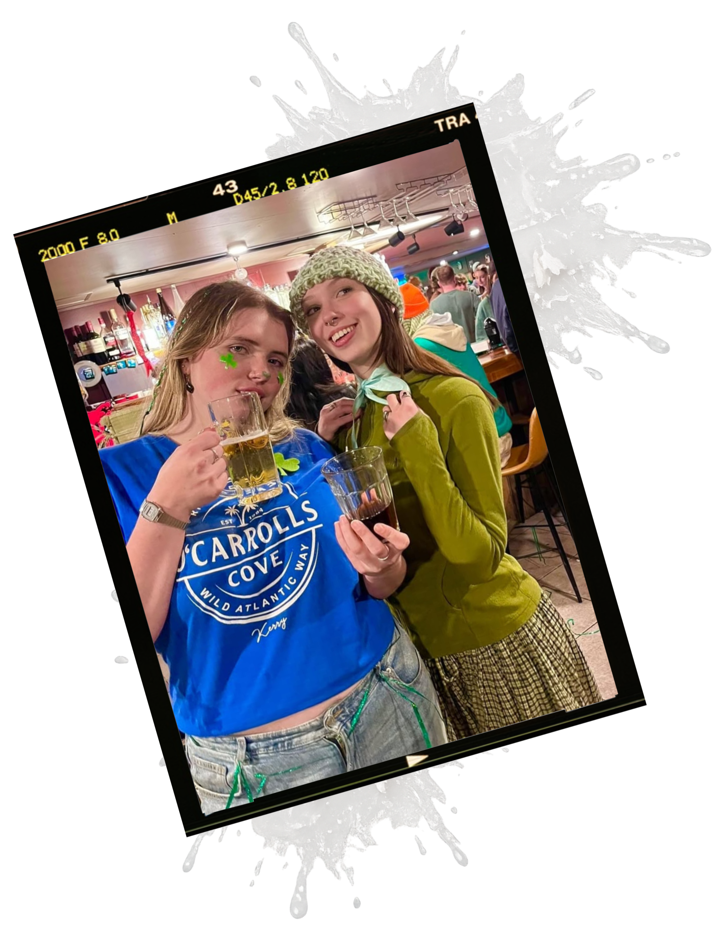 Two young women with painted shamrocks on their faces enjoying drinks at a bar or pub, celebrating during St. Patrick's Day.
