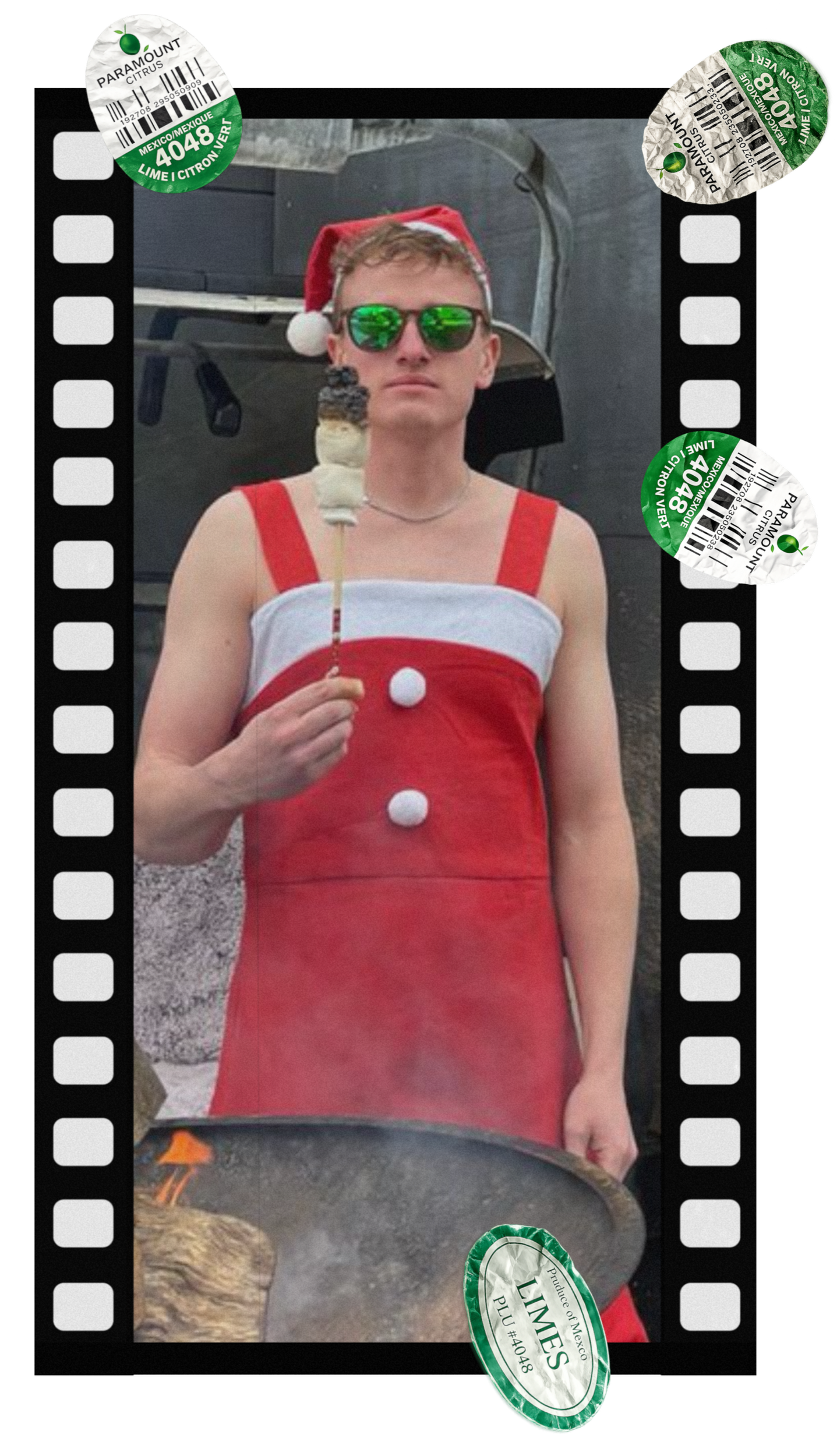 A person dressed as Santa Claus, wearing a red dress with white buttons, green reflective sunglasses, and a red Santa hat with white trim and pom-pom, eating a s'more. They are standing outdoors in front of a grill, with a tongue of flame visible inside the grill.