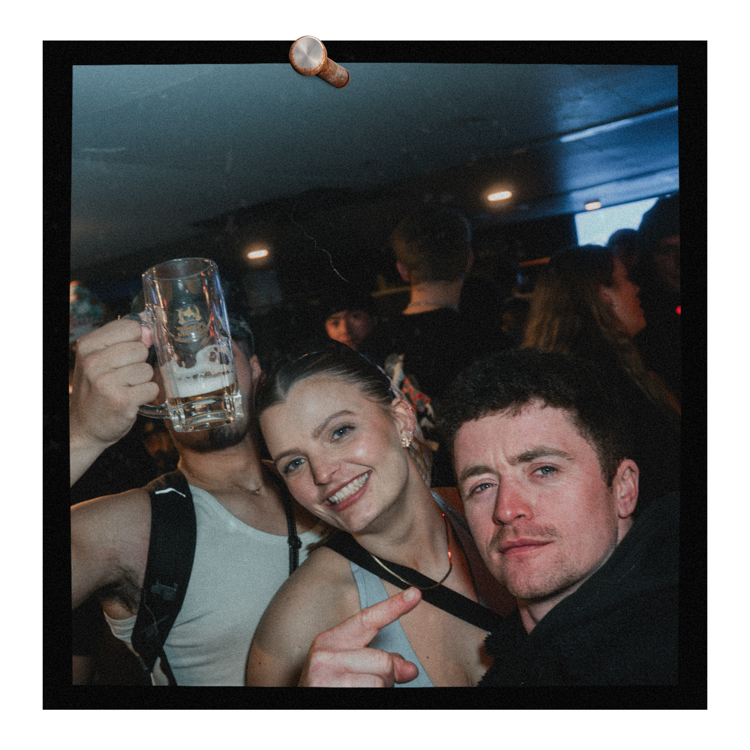 People at a social gathering, with a woman smiling in the foreground and a man making a peace sign in the background, holding a beer mug, in a dimly lit setting.