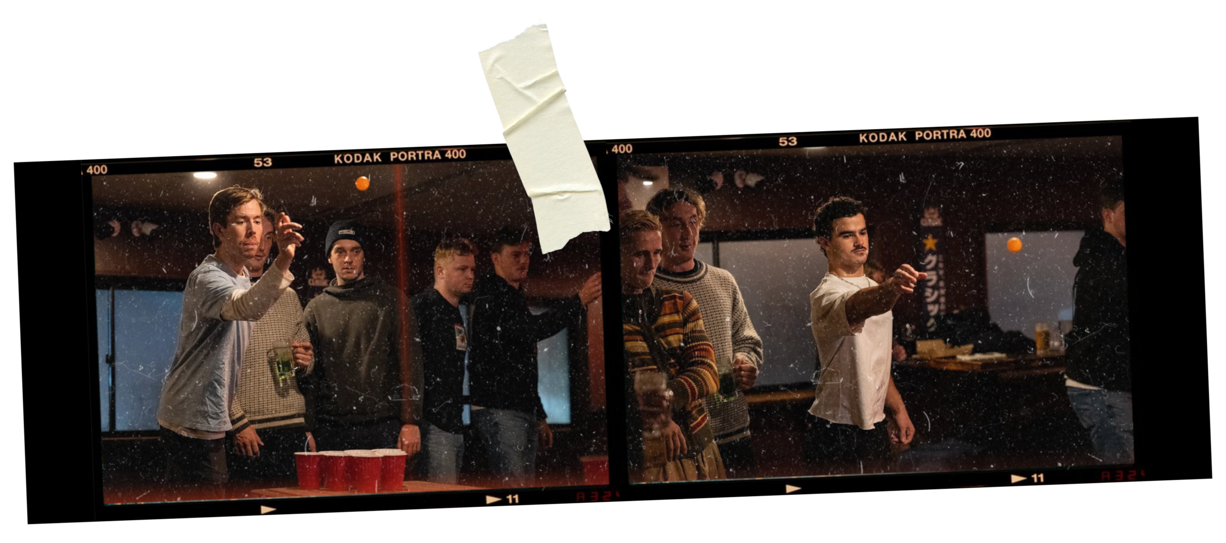 Group of young men playing beer pong inside a dimly lit room, some holding drinks, with a visible reflected scene through a glass panel.