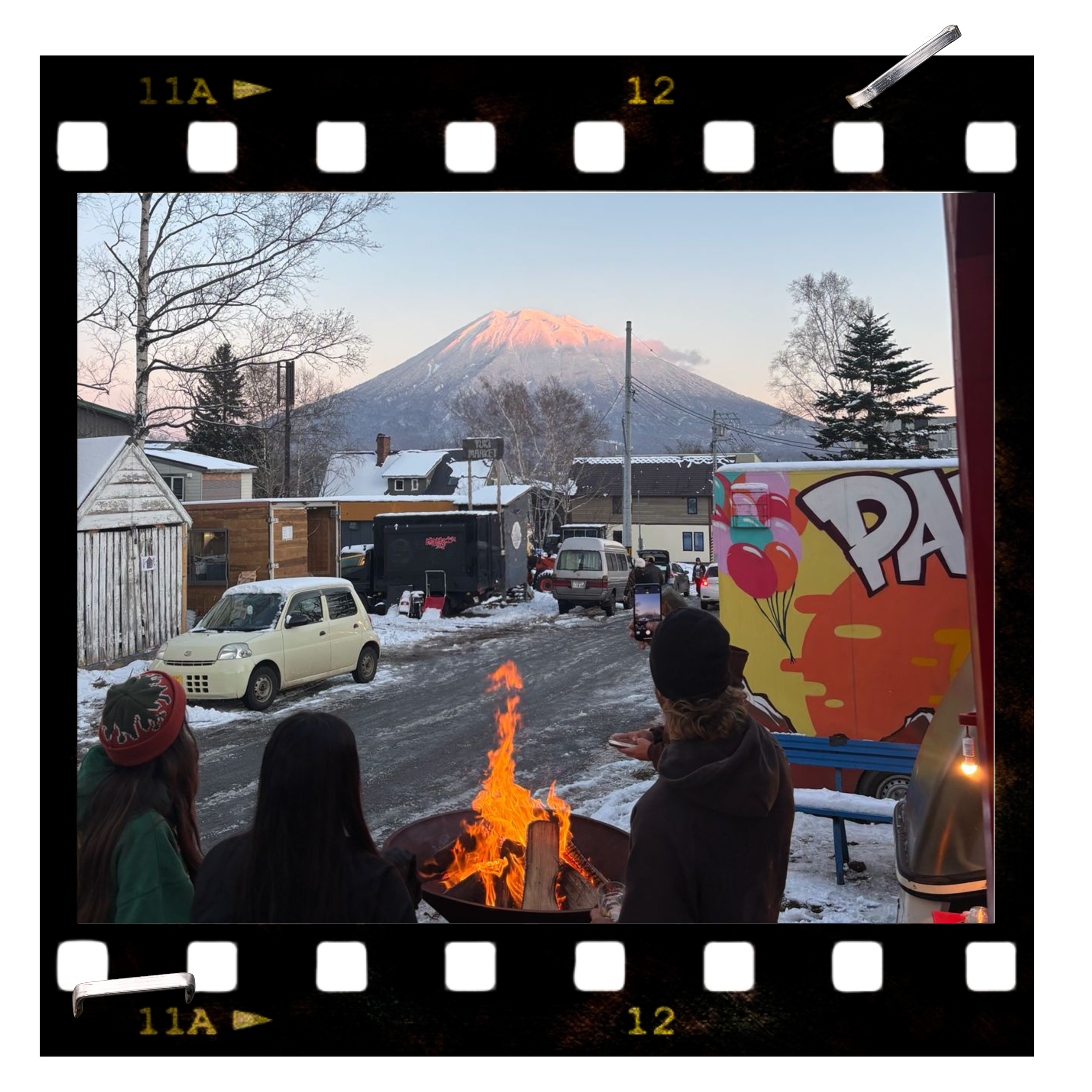 People gathered around a fire outdoors with snow, taking photos of a mountain with a snowy peak at sunset, in front of a sign with a colorful design.