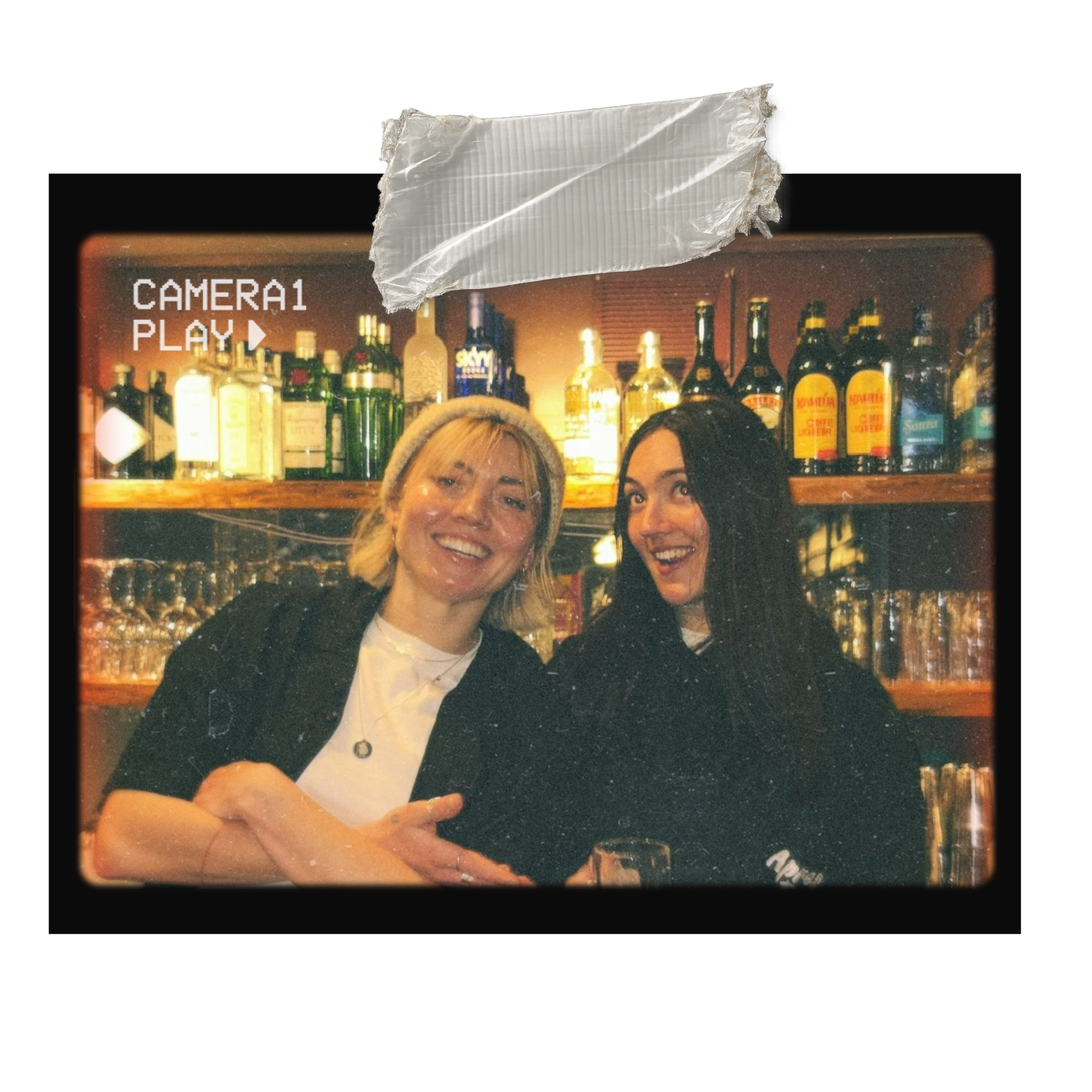 Two women taking a selfie in front of a bar with liquor bottles on display.