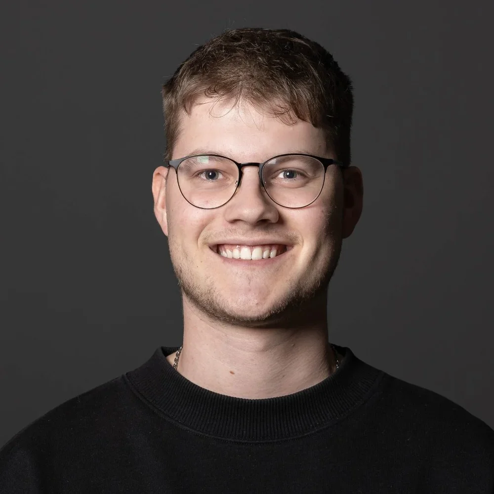Ein junger Mann mit Brille, grauem Hintergrund, trägt einen schwarzen Pullover, lächelt freundlich.
