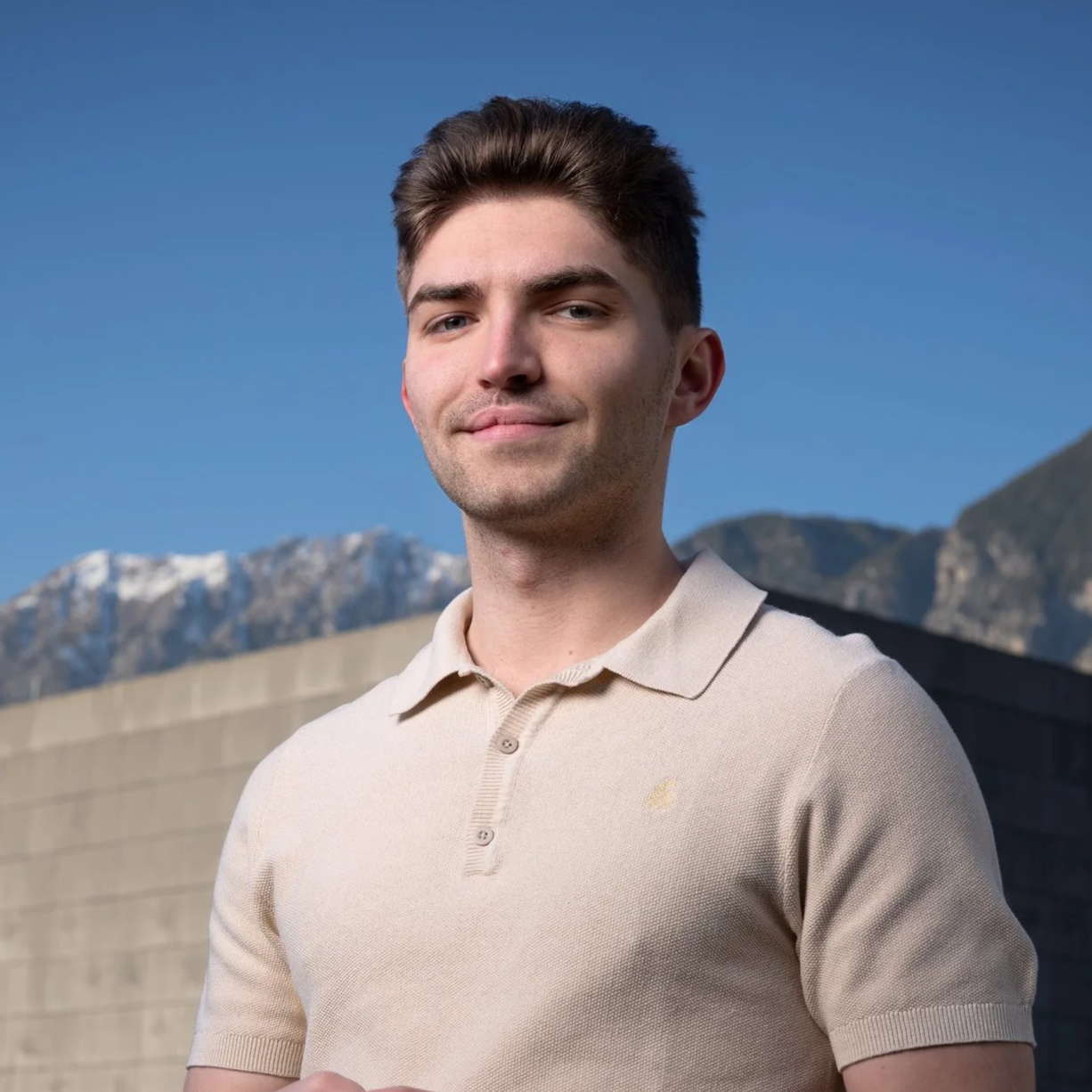 Ein junger Mann mit braunen Haaren, trägt ein beiges Polo-Shirt, steht vor Bergen und blauen Himmel.