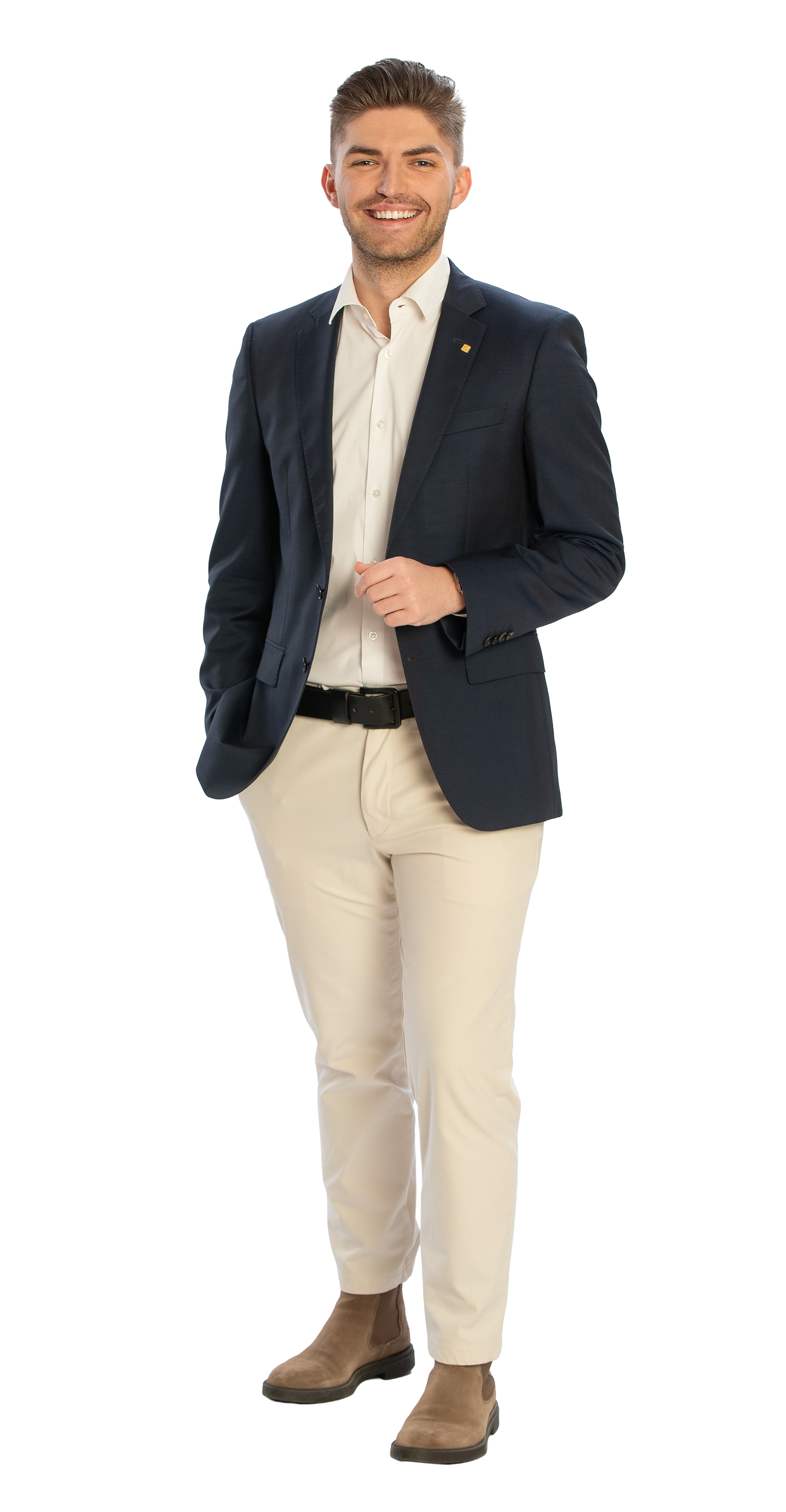 Ein lächelnder Mann in formeller Kleidung, mit dunklem Blazer, weißen Hemd, hellen Hosen, und braunen Stiefeln, steht vor weißem Hintergrund.