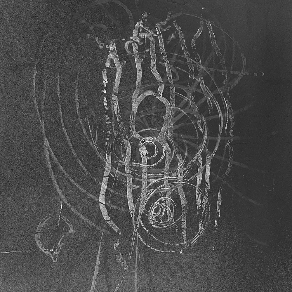A dark photographic surface marked with light scratches and concentric curved forms — Transparency, the physical materiality of the image laid bare.