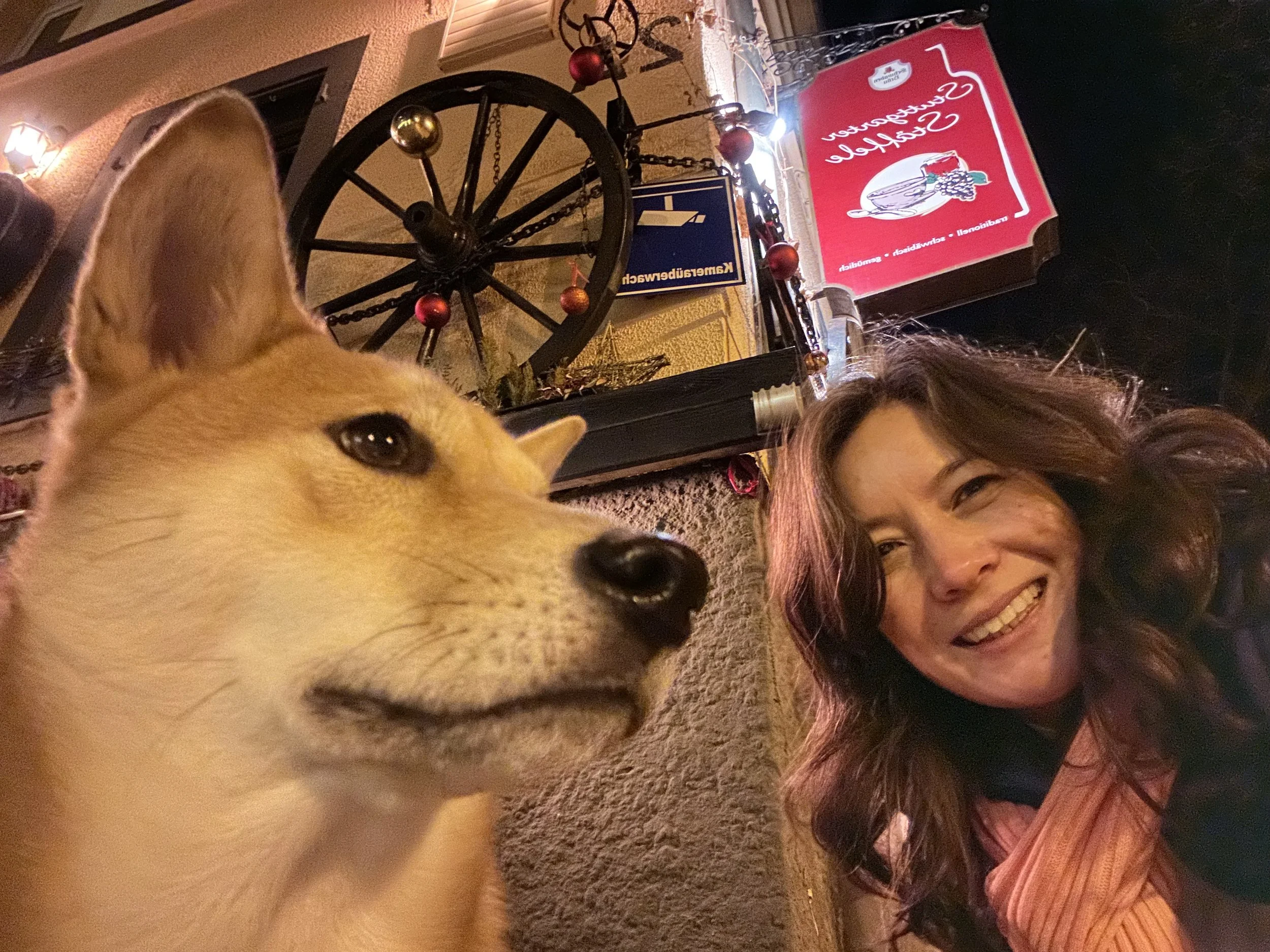A woman smiling with wavy brown hair, wearing a pink top, taking a selfie outdoors at night with a tan and white puppy in the foreground. The background includes a decorated wagon wheel with red ornaments, a sign with a Santa face, and a building wall with lights.
