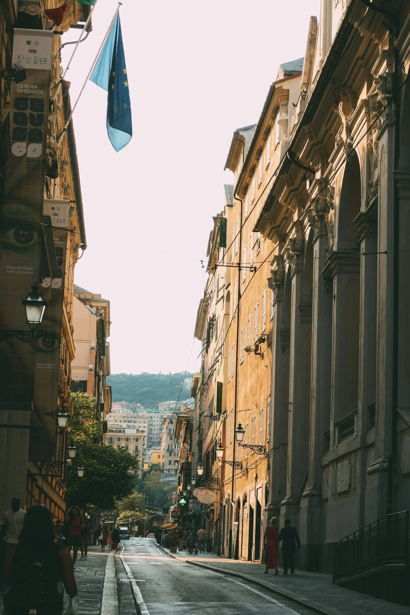 Una strada in un centro storico con edifici antichi su entrambi i lati, alcune persone camminano sul marciapiede e sul lato della strada si notano lampioni e fermate dell'autobus.