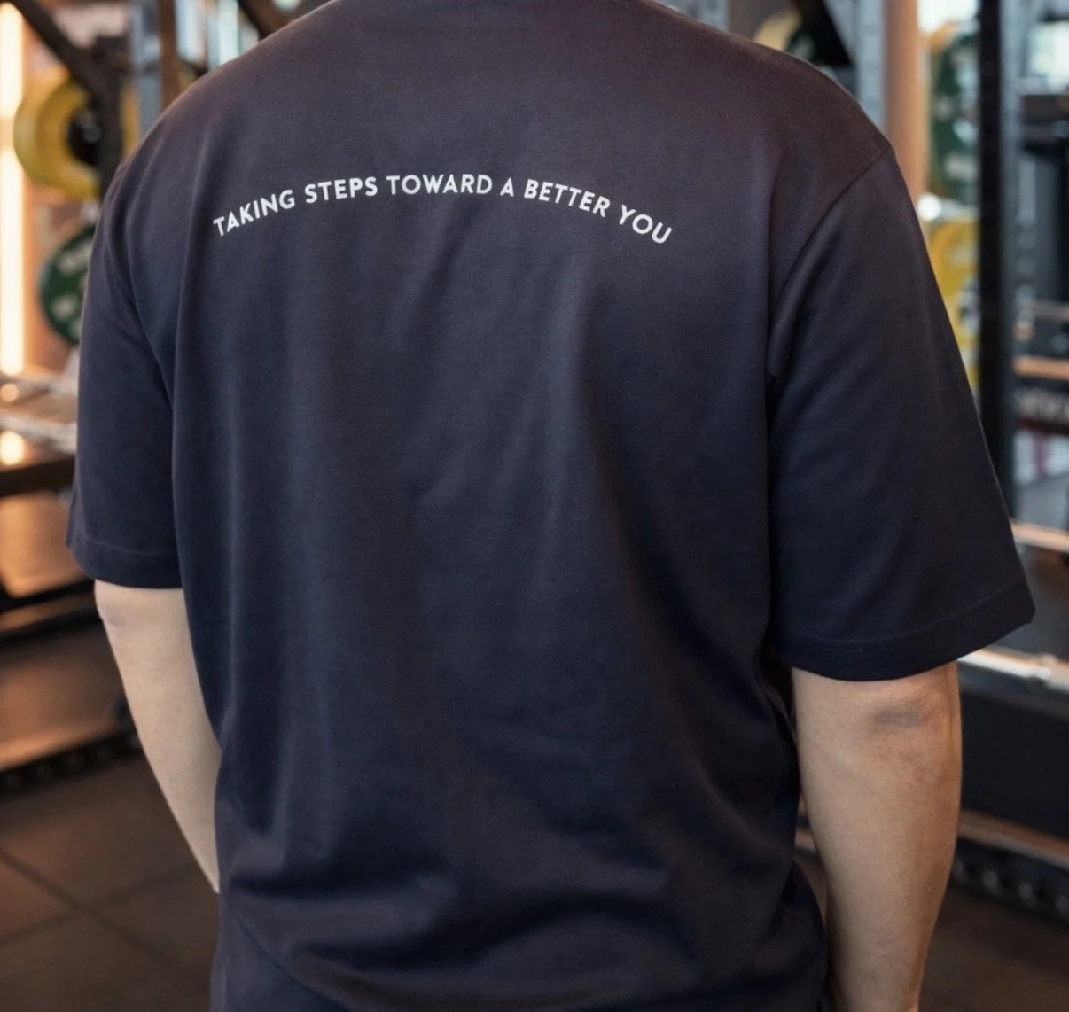 Back of a person wearing a black t-shirt with white text that says 'Taking steps toward a better you' in an indoor setting.