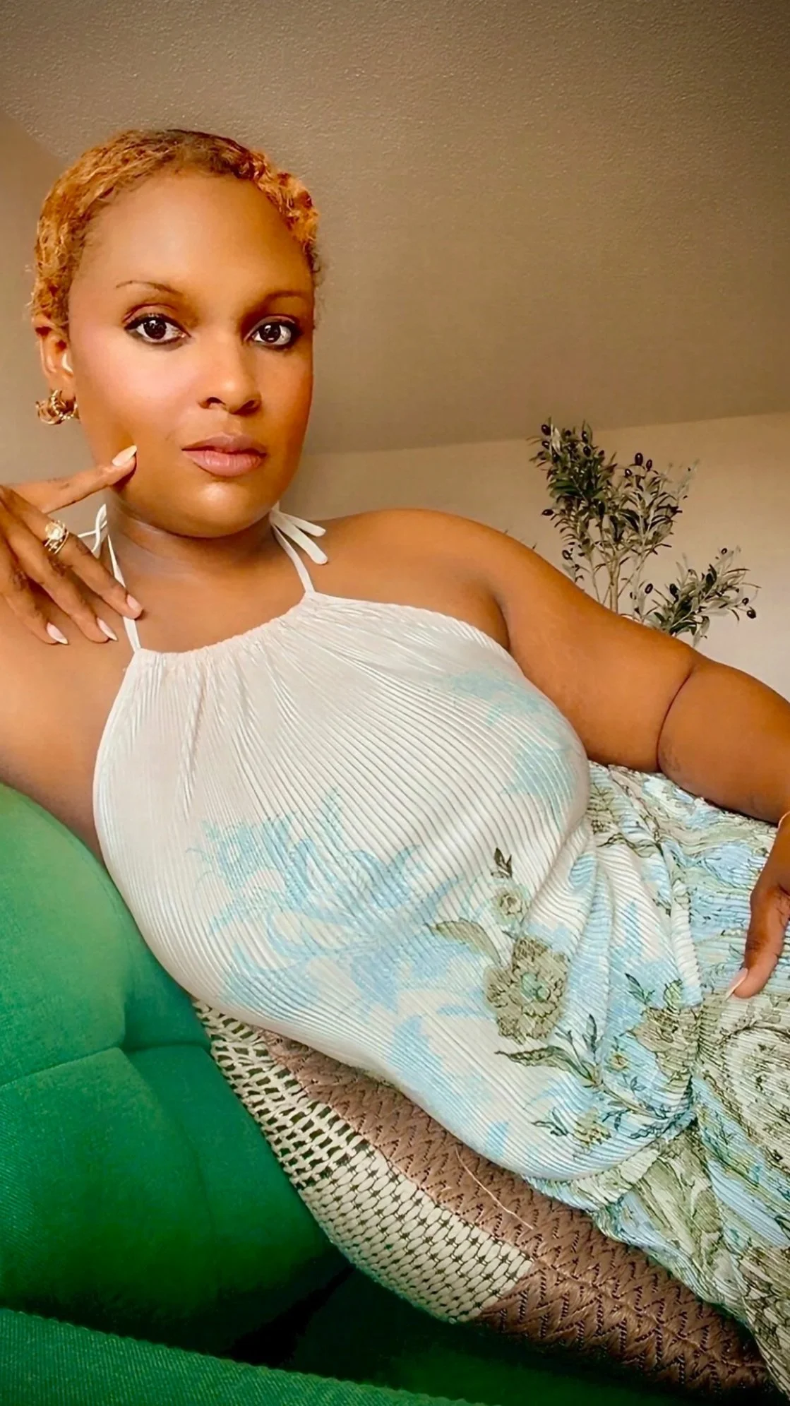 A woman with short curly hair, earrings, and makeup, wearing a sleeveless light-colored dress with floral patterns, sitting on a green bench or chair with a plant in the background.