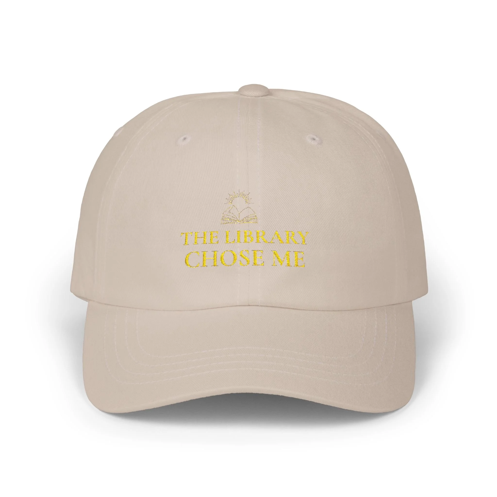 Beige baseball cap with gold embroidered text 'The Library Chose Me' and a small gold embroidered open book with a sun above.