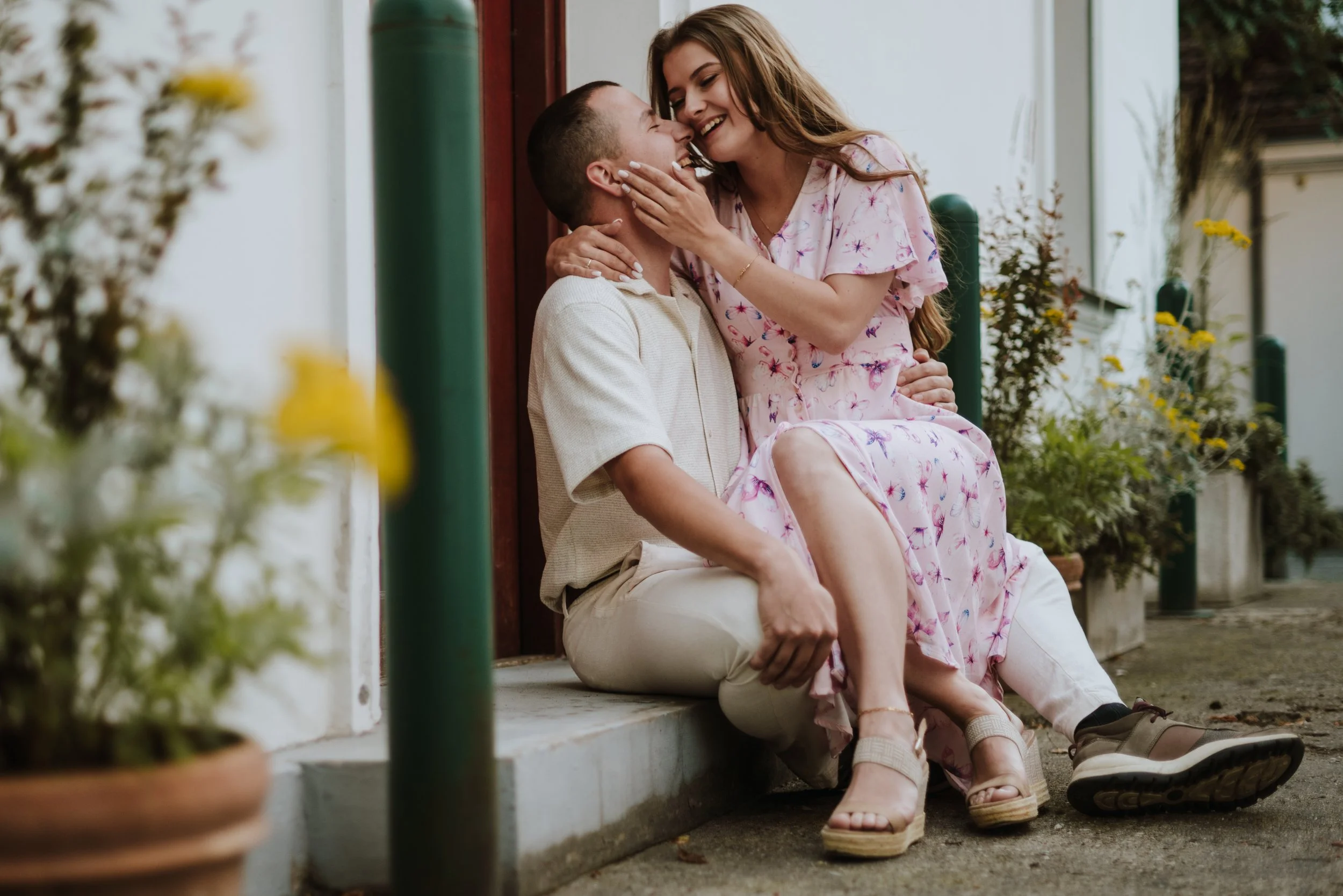 A young couple sitting on a porch, with the woman sitting on the man's lap, sharing a joyful and intimate moment. The woman has long hair and is wearing a pink floral dress, and the man is dressed in light-colored clothing. They are surrounded by pot