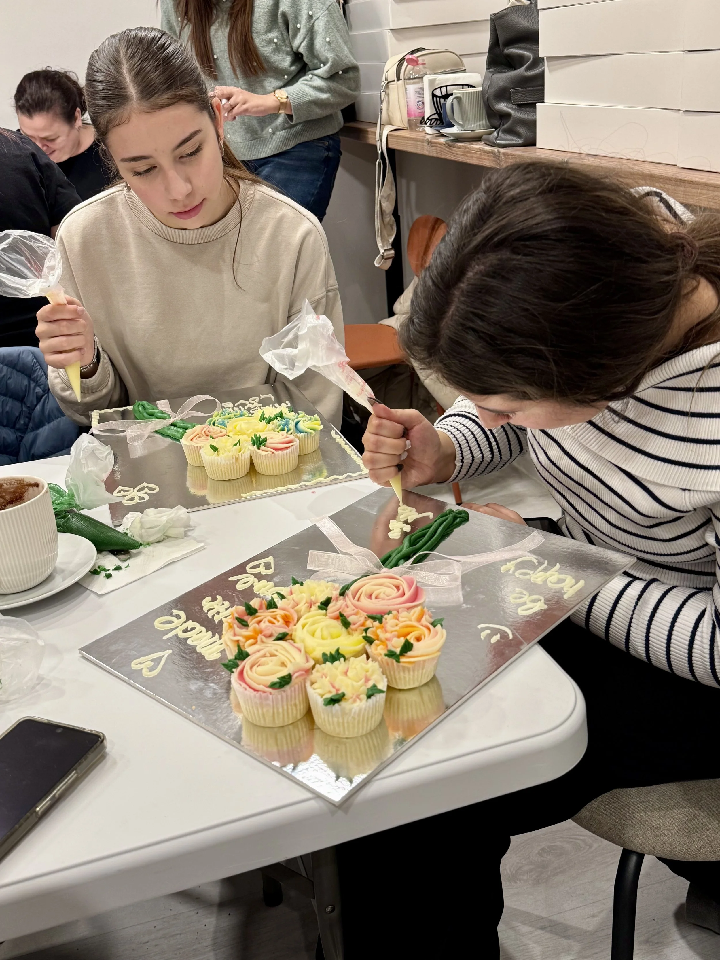 Two women decorating cupcakes with pink and yellow frosting, surrounded by piping bags, cupcakes on a tray, and a cake board with messages and hearts at a gathering.