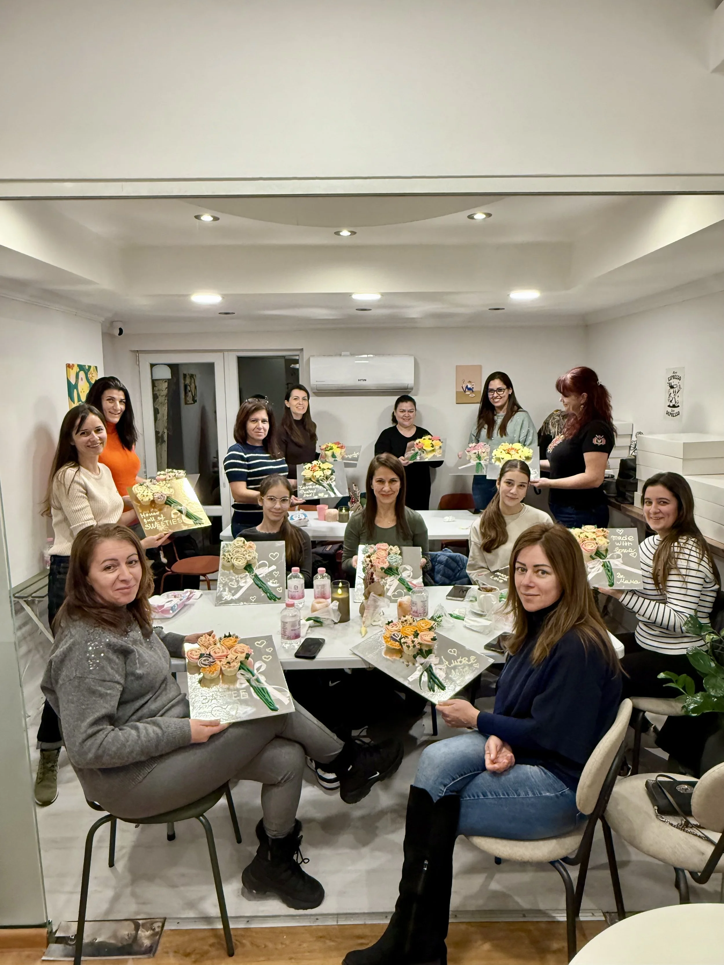 A group of twelve women gathered in a room, holding flower arrangements and decorated boxes, celebrating a special occasion with smiles and flowers.