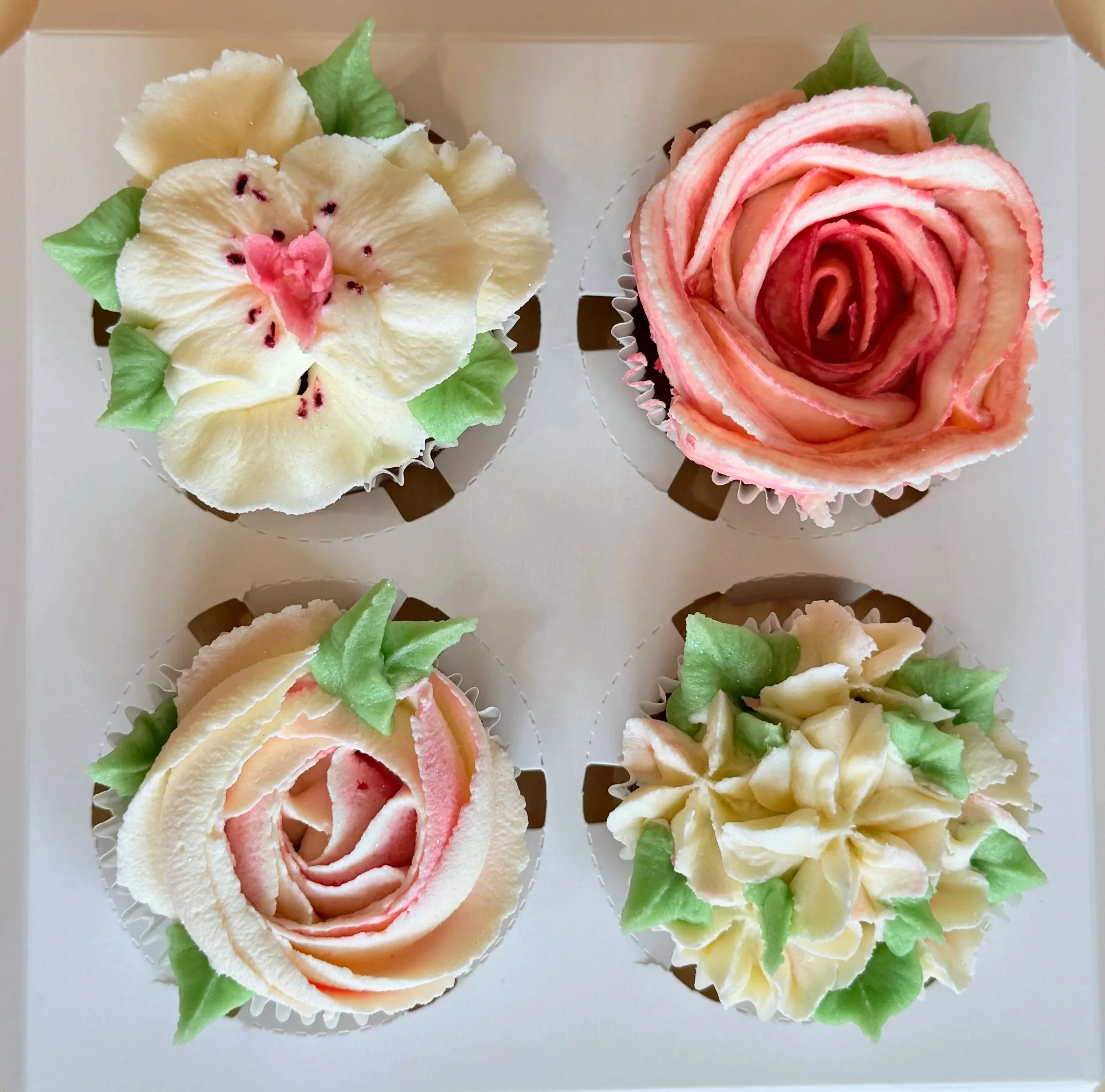 Four cupcakes decorated with flower and leaf designs, arranged in a white box.