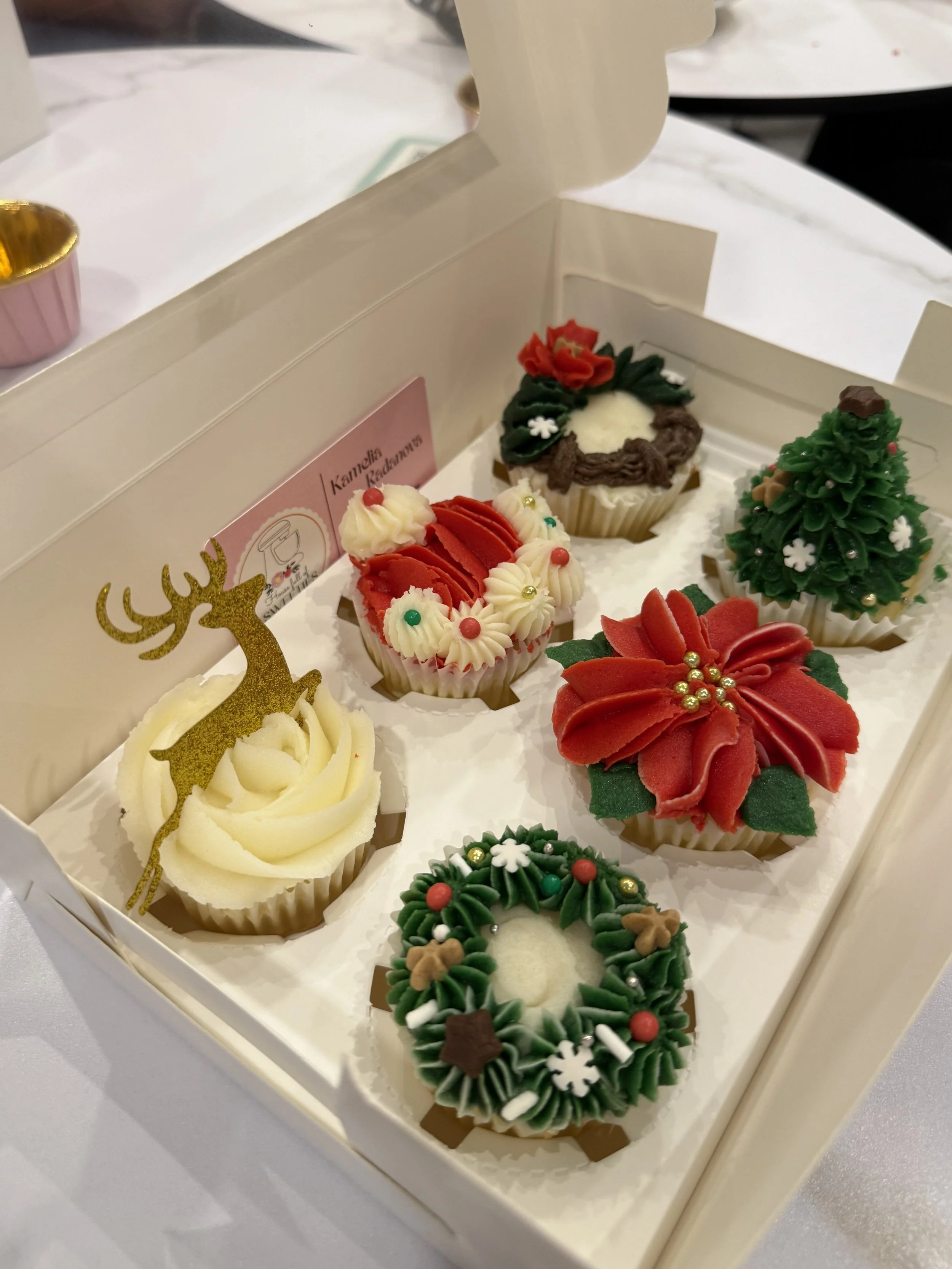 Christmas-themed cupcakes in a white box, decorated with poinsettia, holly, Christmas tree, snowflake, and reindeer motifs.