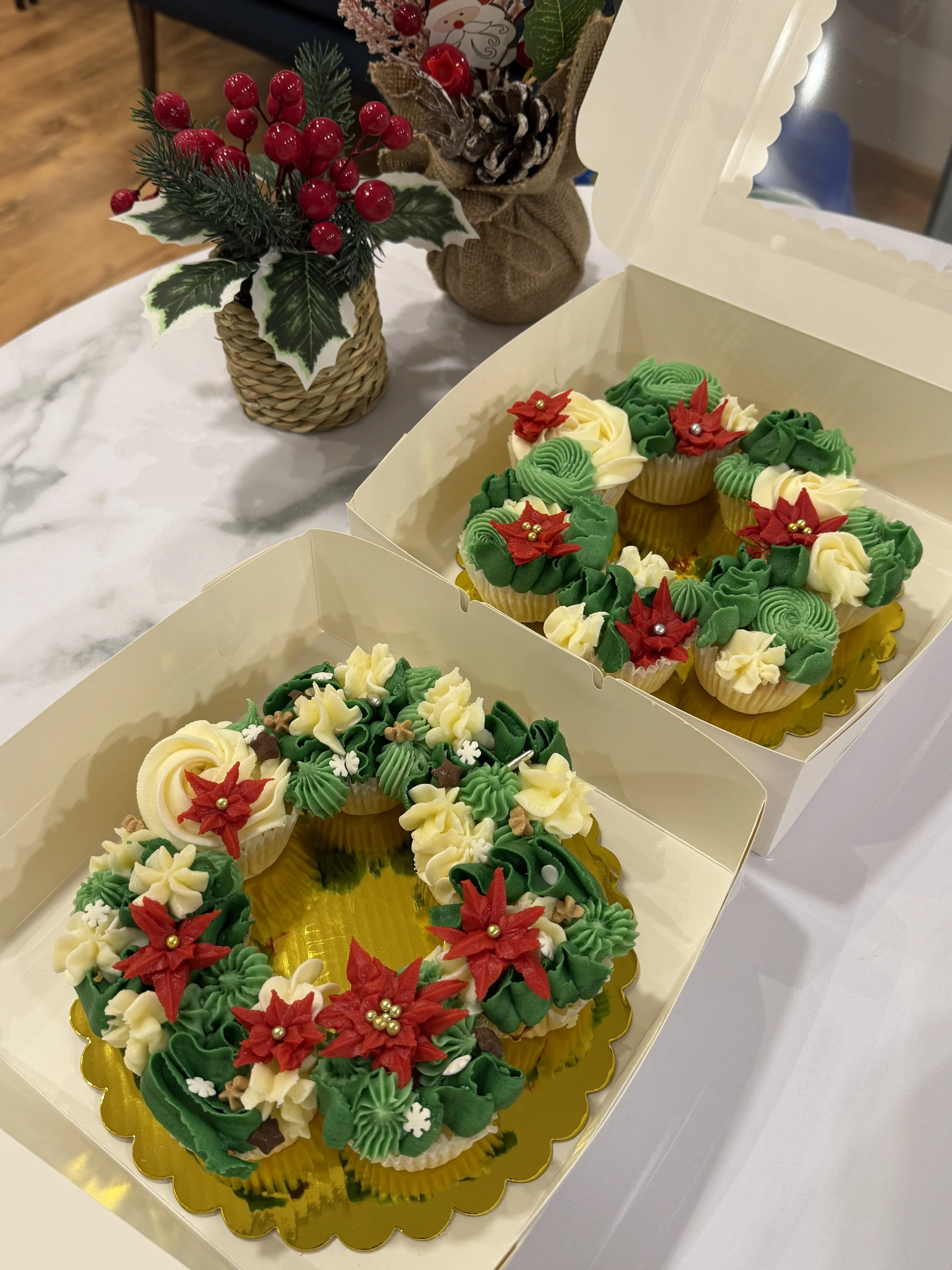 Cupcakes decorated with Christmas-themed green, red, and white frosting and poinsettia design, in a festive box on a white table with holiday decorations in background.