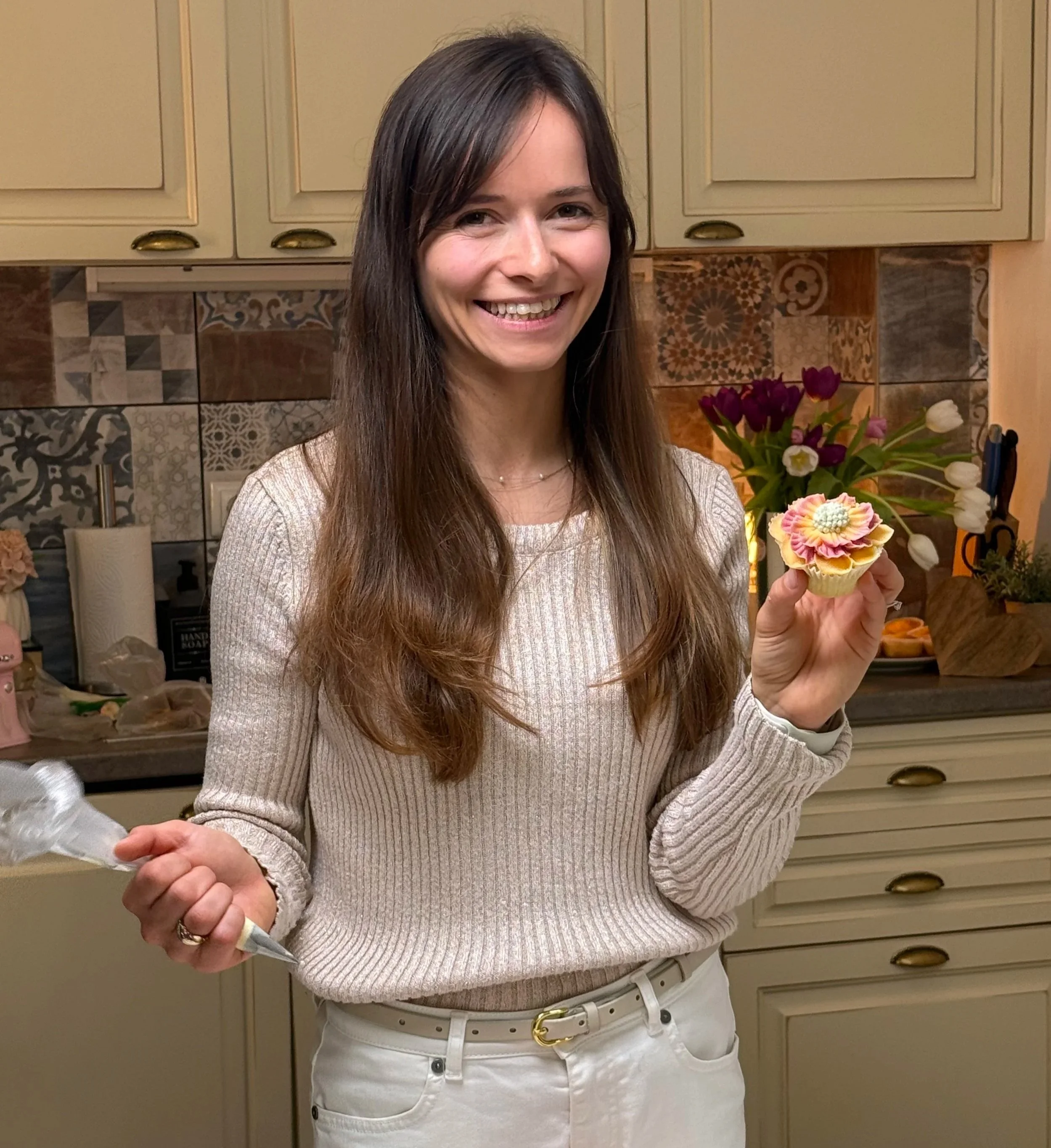A woman with long brown hair and a beige sweater holding a decorated cupcake, smiling in a kitchen with a floral bouquet and patterned tile backsplash.