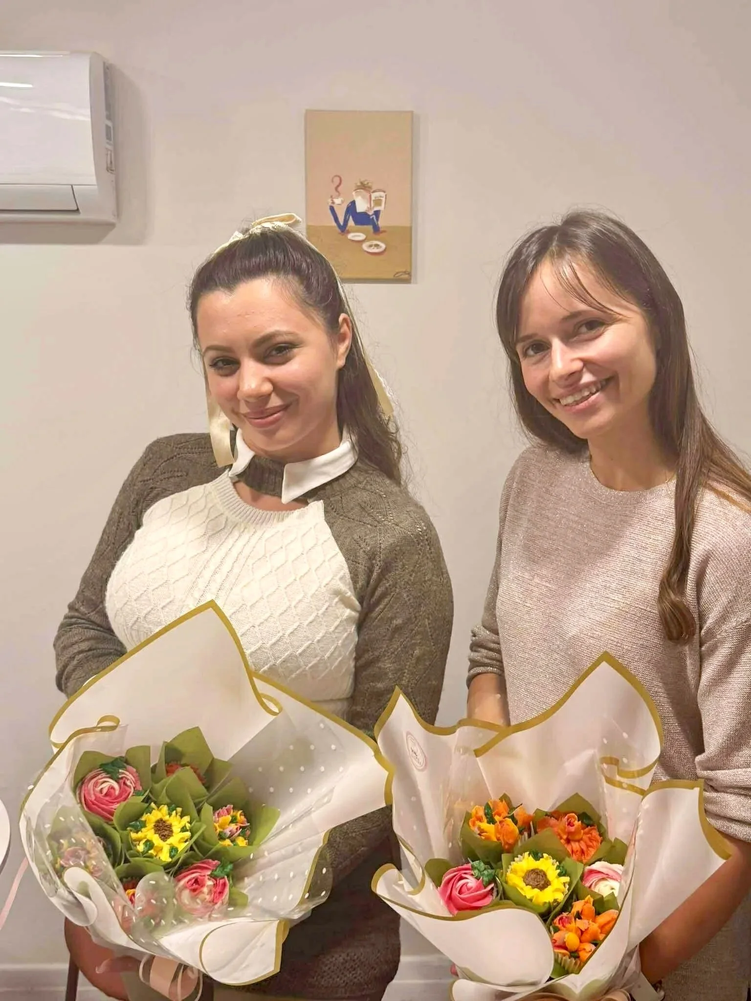 Two women standing indoors, holding bouquets of colorful flowers, smiling at the camera.