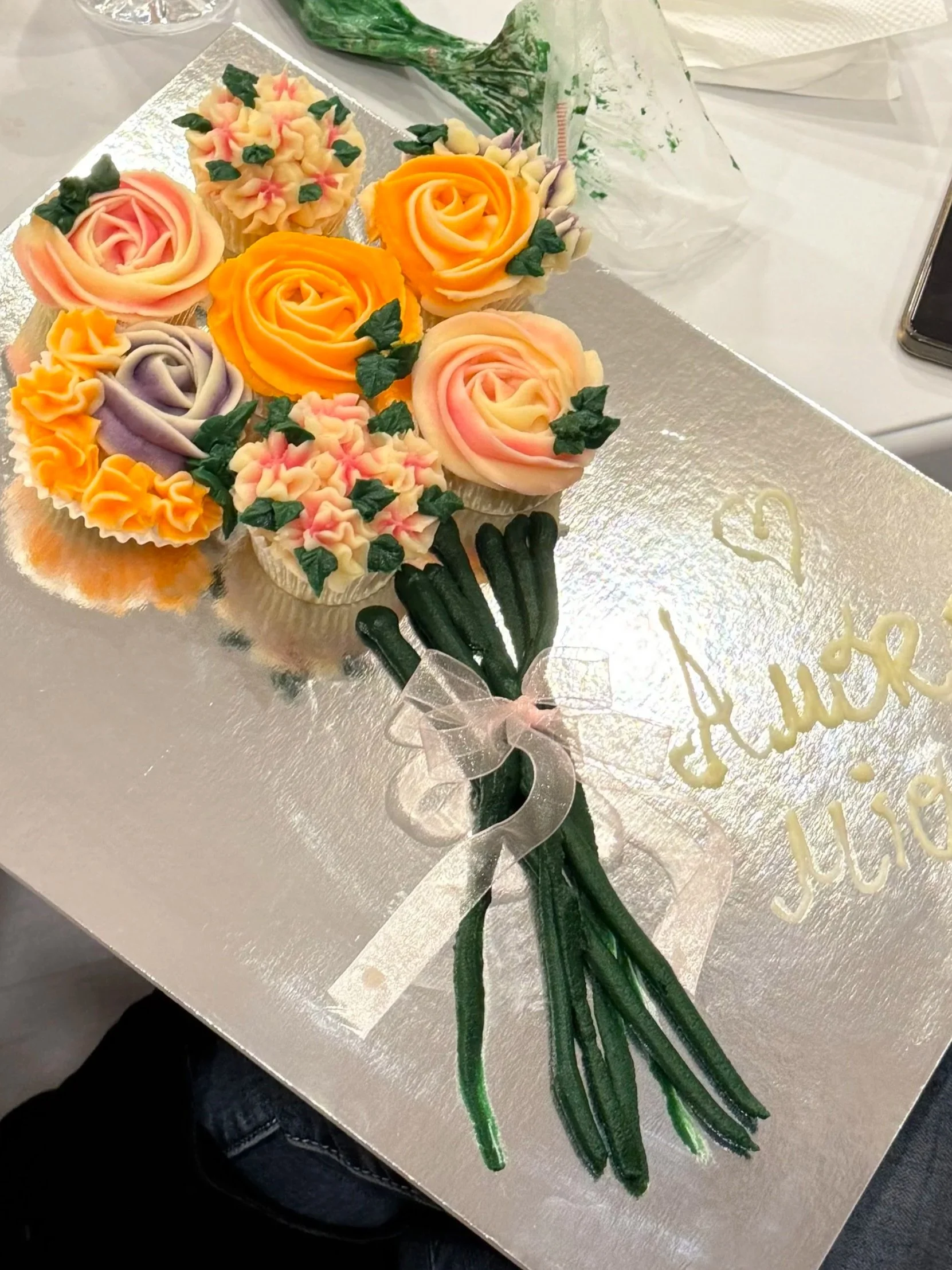 Cake decorated to look like a bouquet of colorful roses with green leaves, arranged on a silver cake board with gold writing.