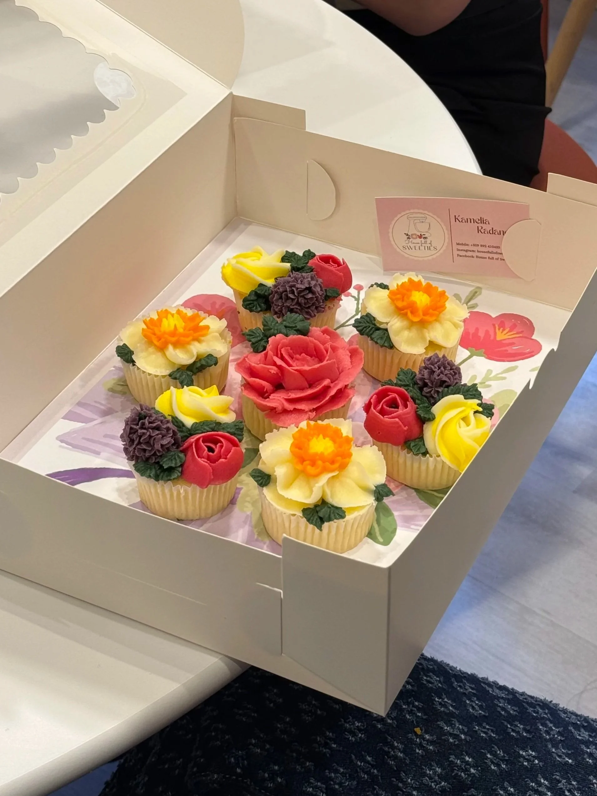 A white box containing decorated cupcakes designed to look like colorful flowers, including roses and daffodils, with a business card inside the box.