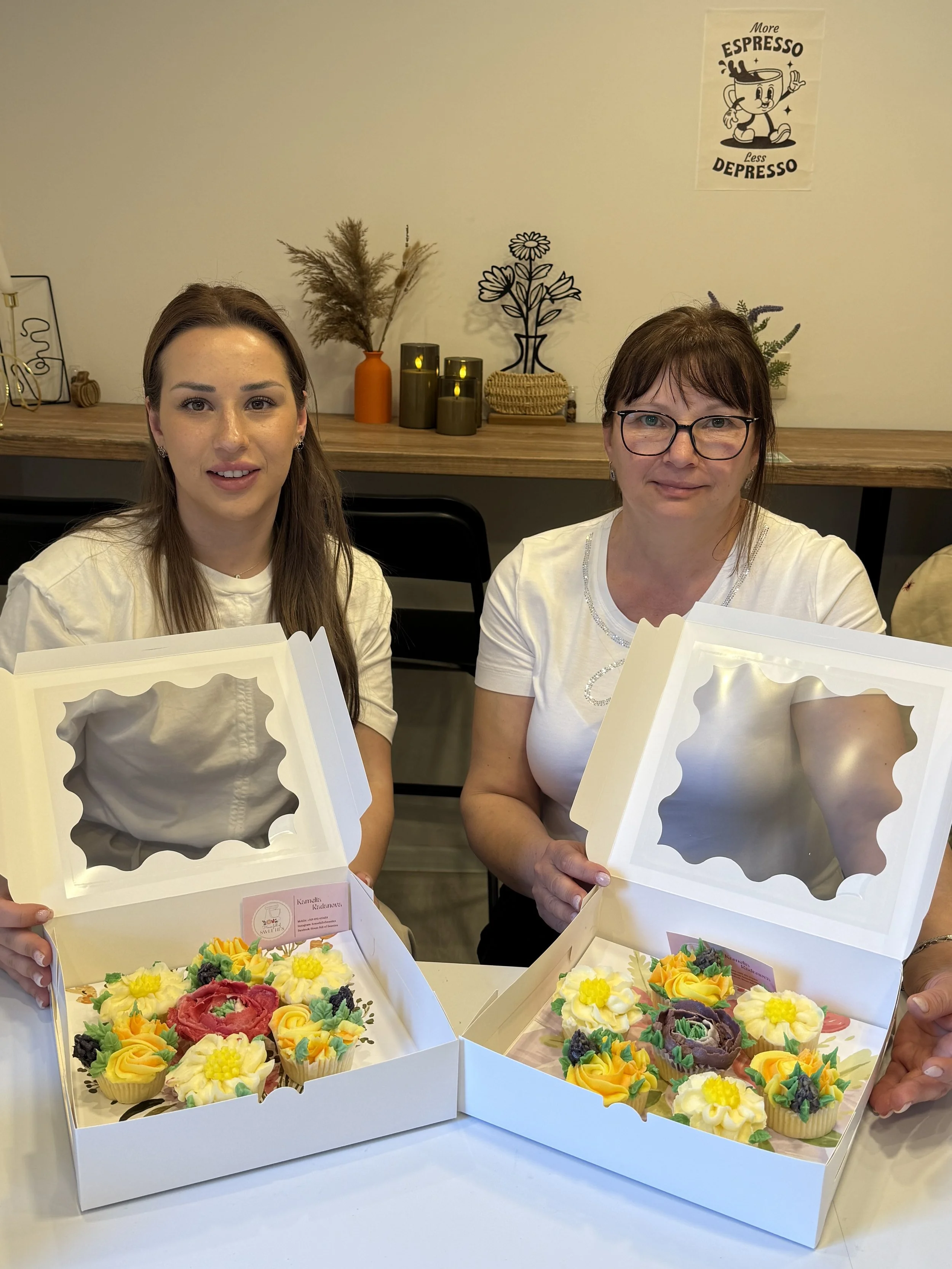 Two women sit at a table holding open boxes of decorated cupcakes with colorful frosting. One woman has long brown hair, and the other has short brown hair and glasses. A wall with decorative plants, candles, and a poster is in the background.