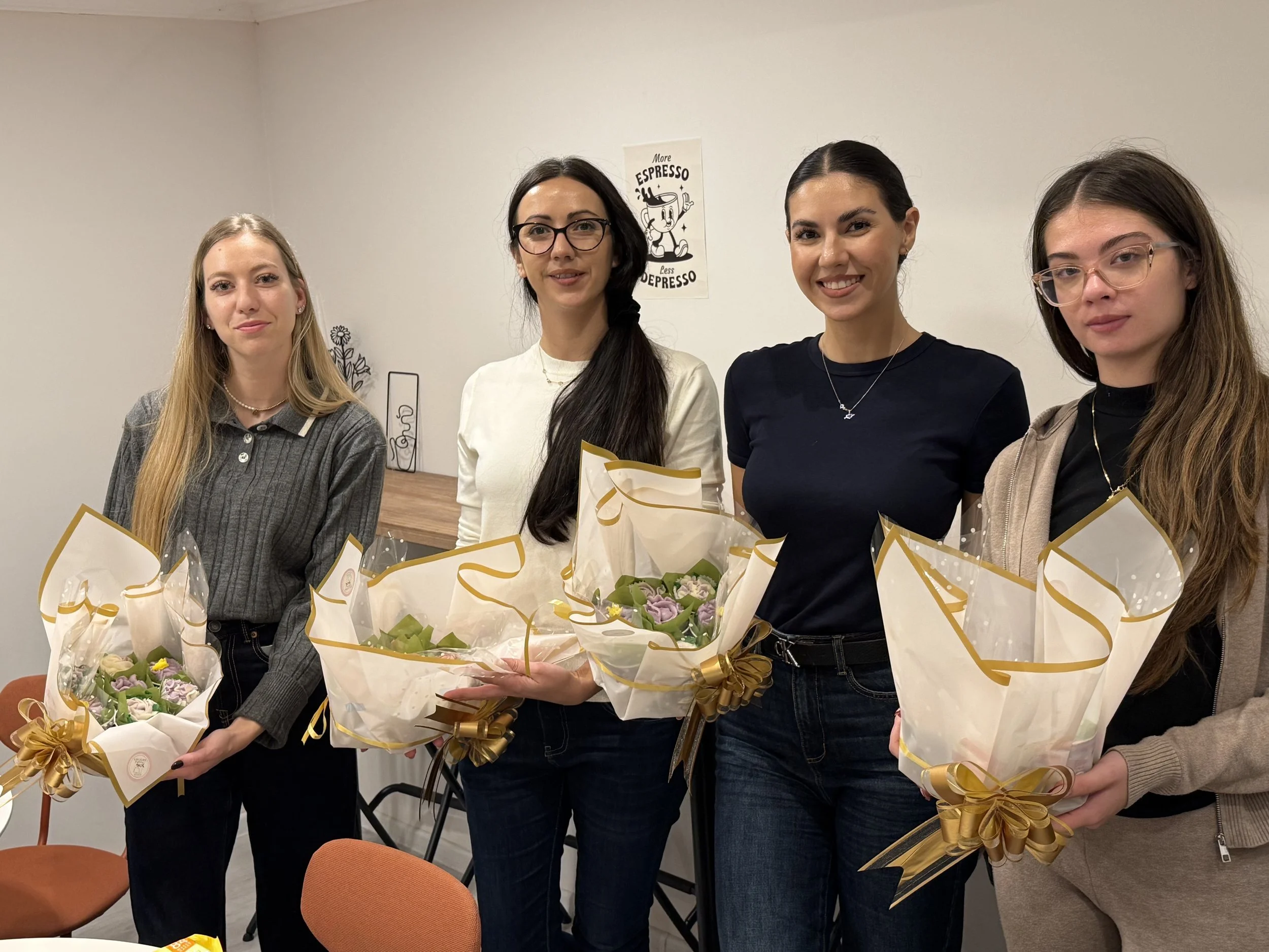 Four women standing in a row, each holding a bouquet of flowers, in an indoor setting with white walls and a small wooden table nearby.