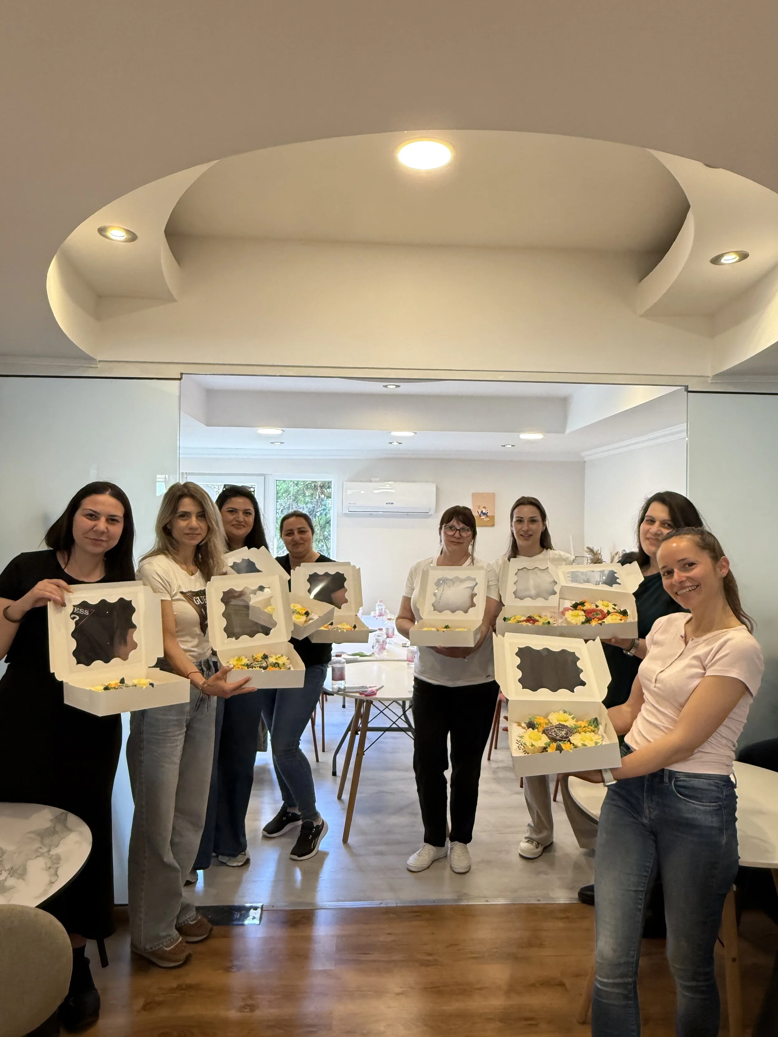 A group of women stands together in a room, each holding a pizza box with a decorated pizza inside. They are smiling and appear to be celebrating or participating in a gathering.