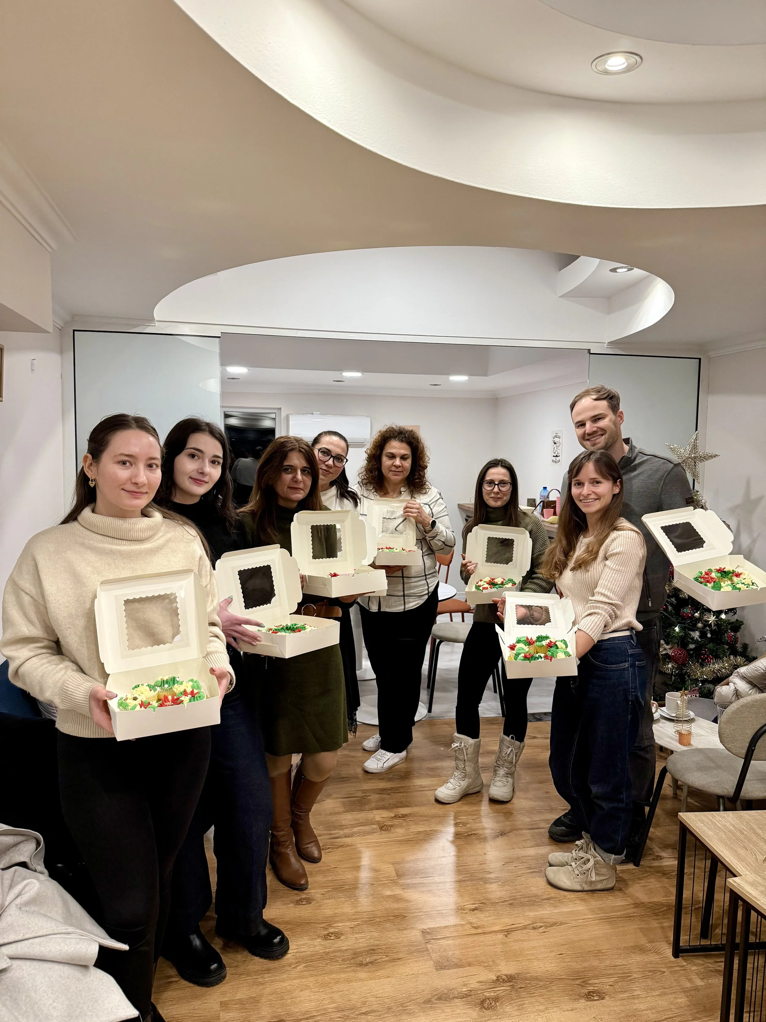 Group of seven women and one man holding decorated holiday cakes in gift boxes during a holiday gathering in a living room with a Christmas tree in the background.