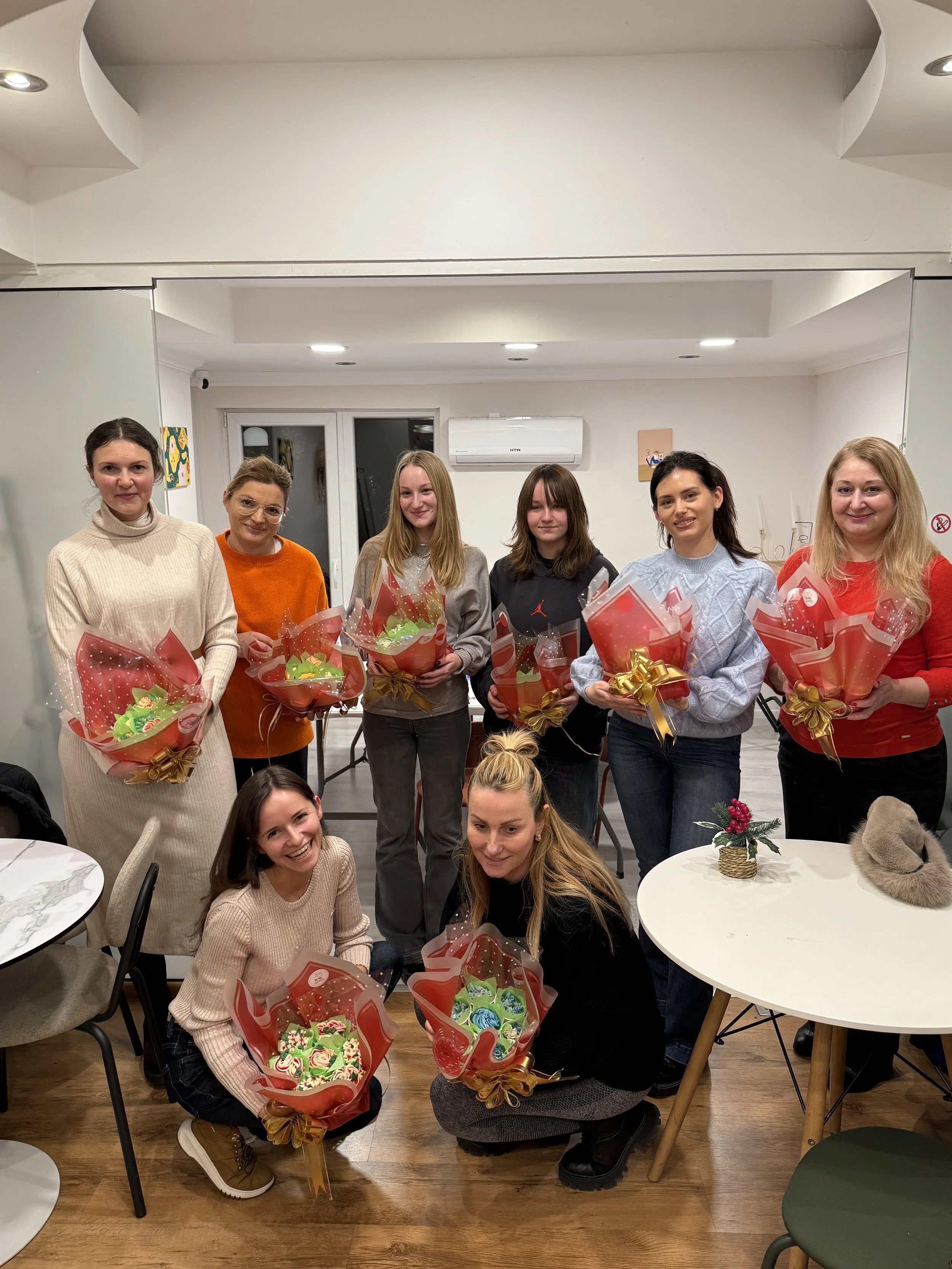 Ten women holding gift baskets with flowers, smiling, gathered in a well-lit indoor room with tables and chairs.
