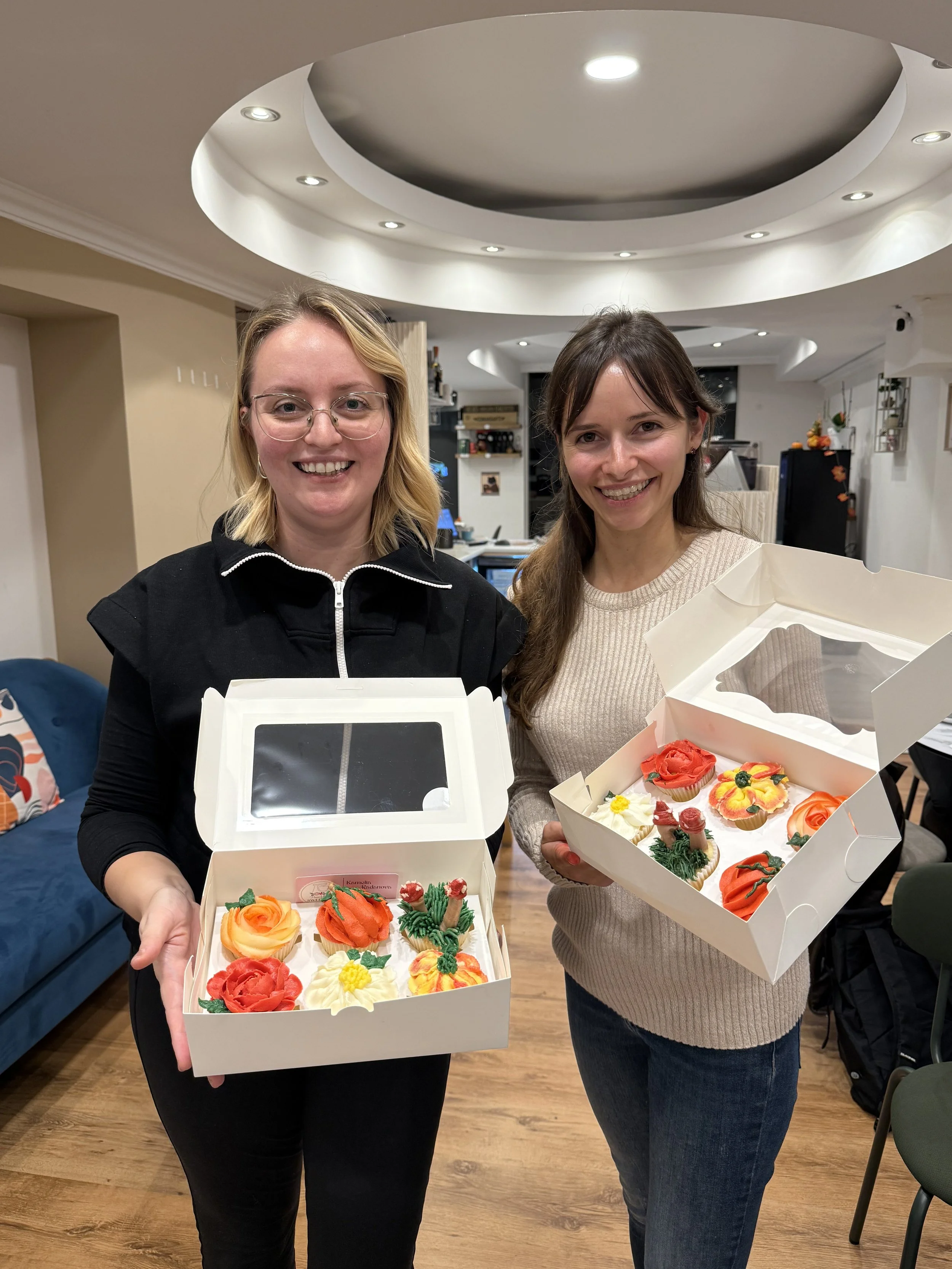 Two women smiling and holding boxes of decorated cupcakes in a cozy room.