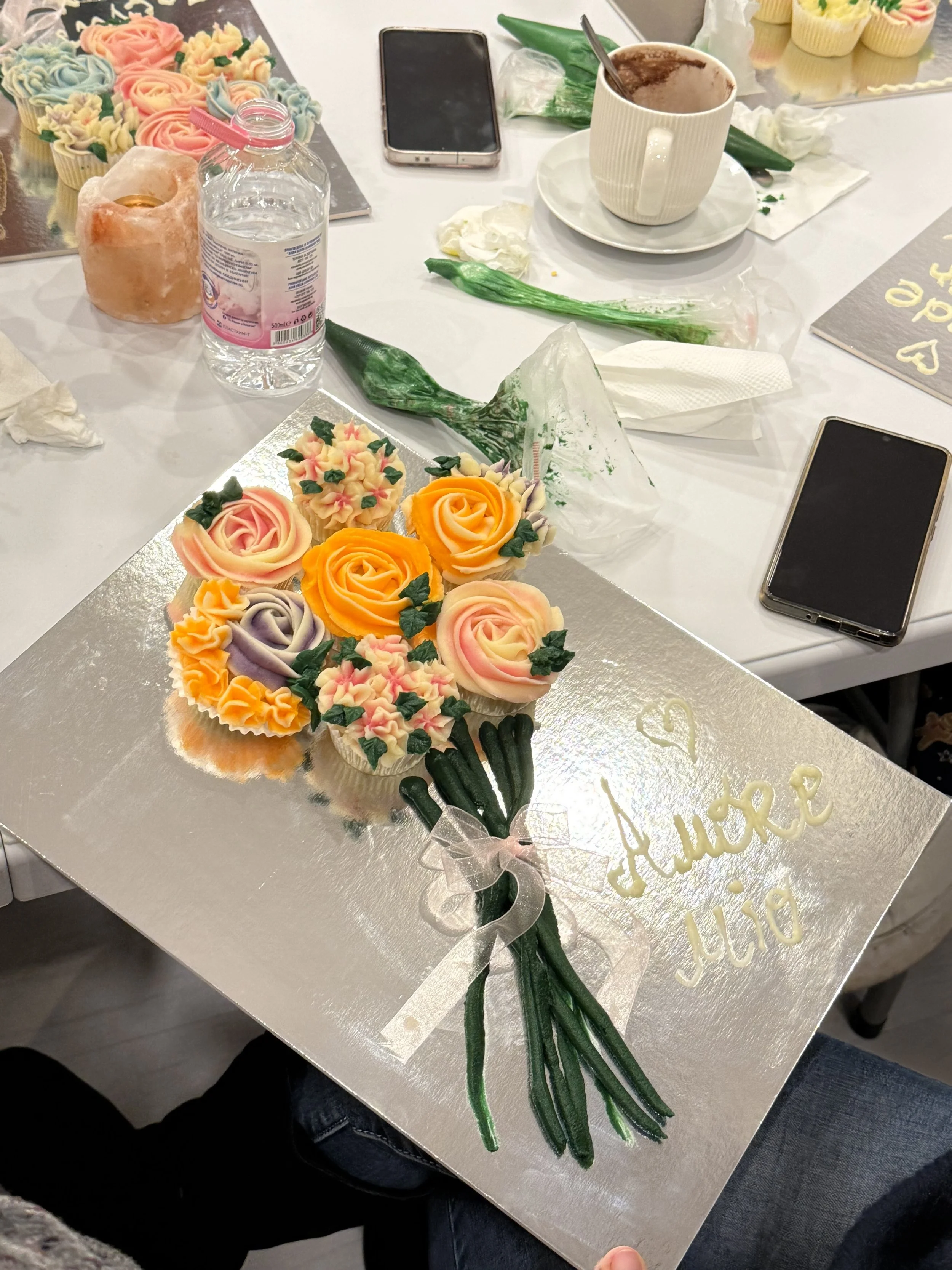 A bouquet of colorful roses and flowers made of buttercream or similar edible material, laying on a silver cake board. The bouquet contains peach, purple, yellow, and pink roses with green leaves and small pink flowers. There are also some green flower stalks with buds attached, and the bouquet is tied with a clear ribbon.