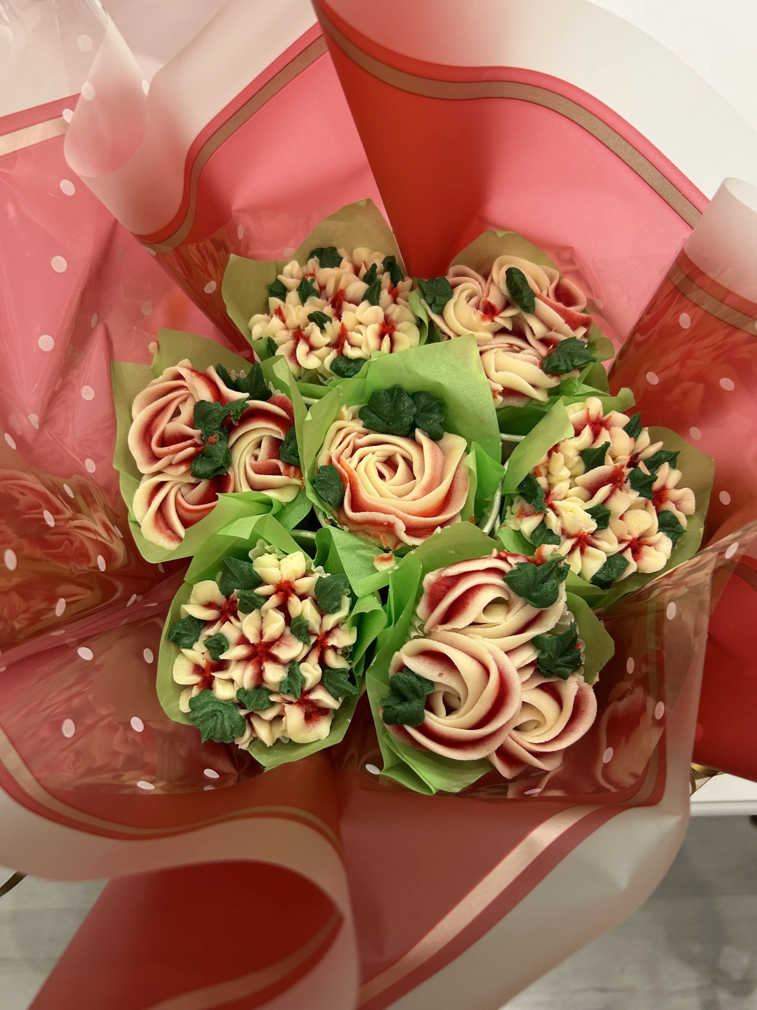 Bouquet of decorative cupcakes with rose and floral designs, wrapped in pink and red polka-dotted cellophane.