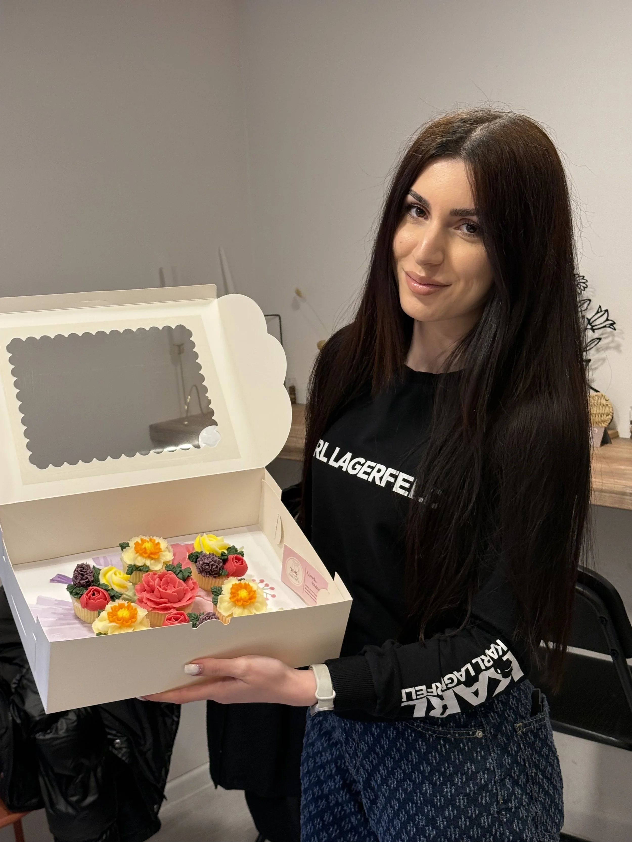 A woman with long dark hair holding a white box decorated with colorful flower-shaped cupcakes or cake decorations, smiling at the camera.