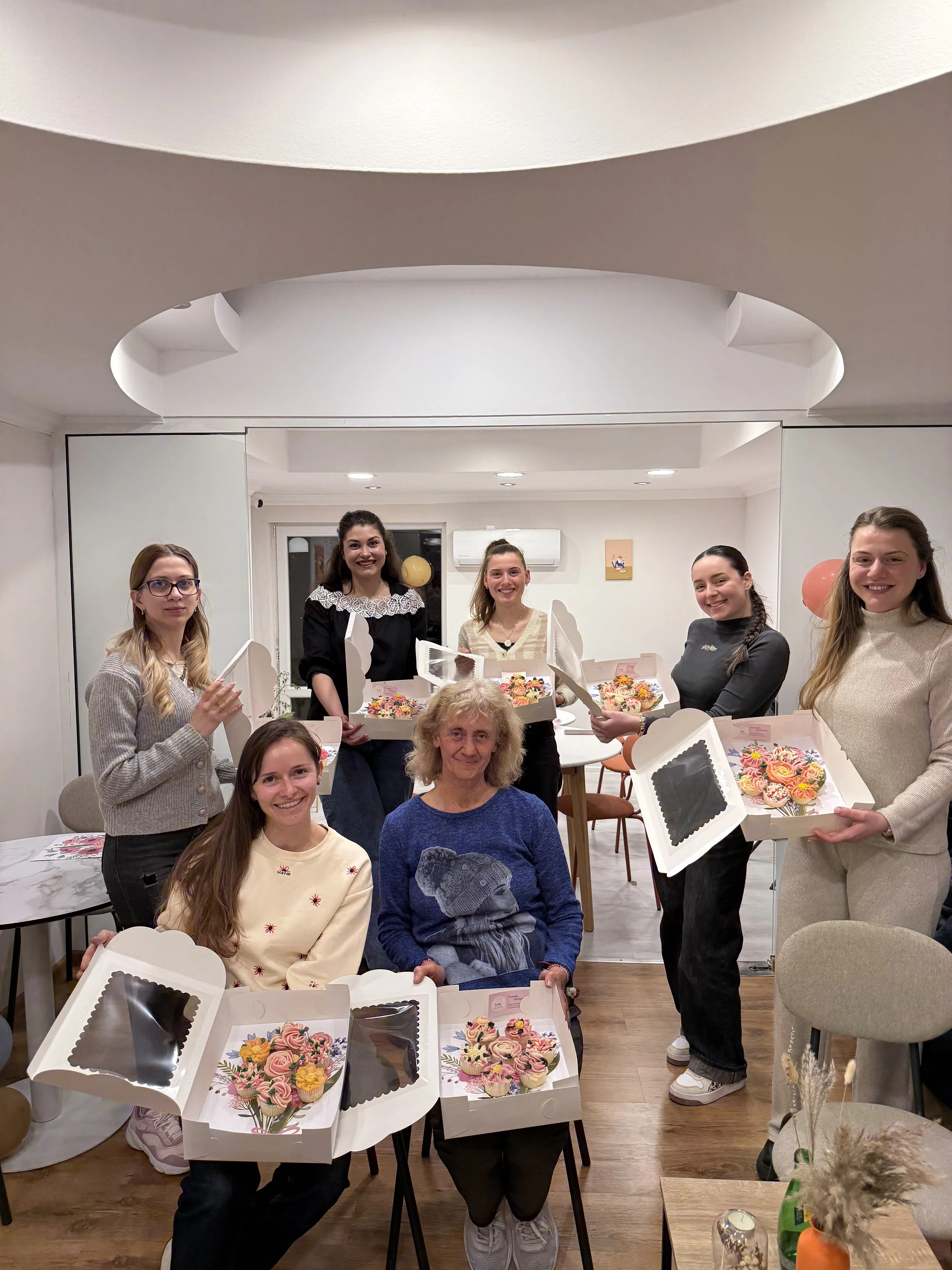 A group of women gathered in a room celebrating, holding boxes of decorated cupcakes, smiling and posing for the photo.
