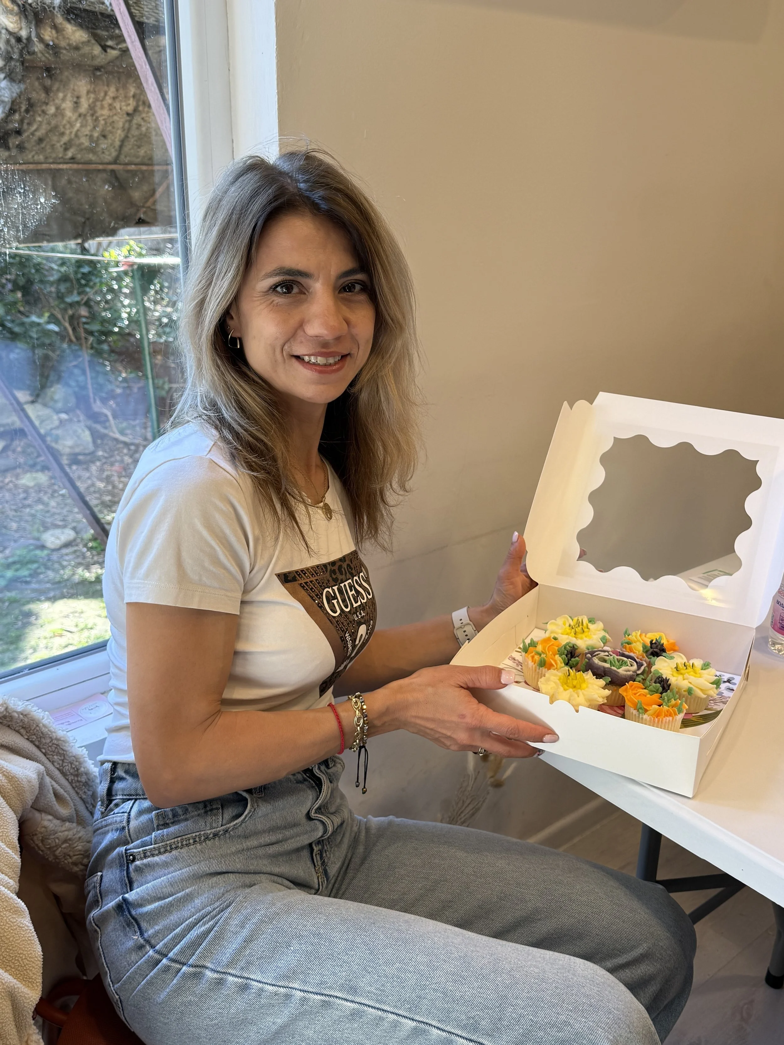 Woman sitting by a window holding a box of colorful cupcakes with yellow, purple, orange, and black icing.