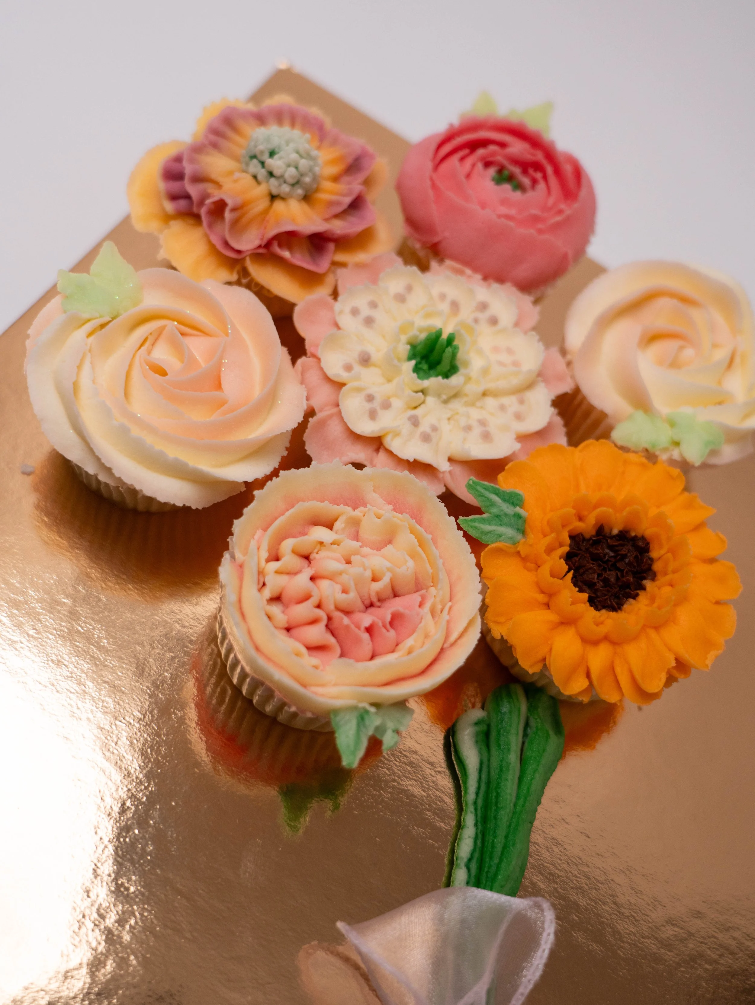 Decorative cupcakes shaped like a bouquet of flowers, including roses, sunflowers, and daisies, on a gold surface.