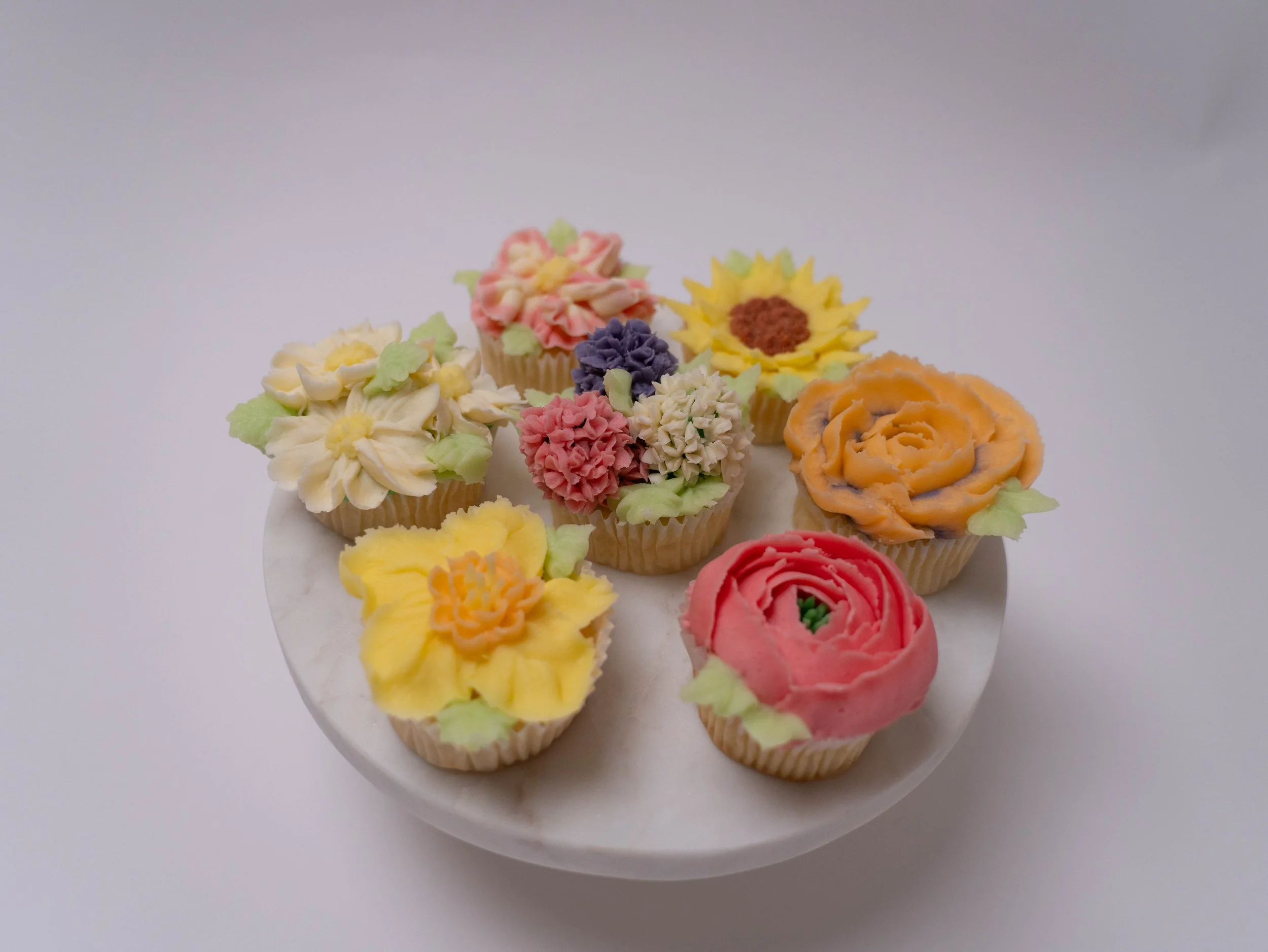 Assorted colorful cupcakes decorated with flower-shaped icing on a white marble plate.